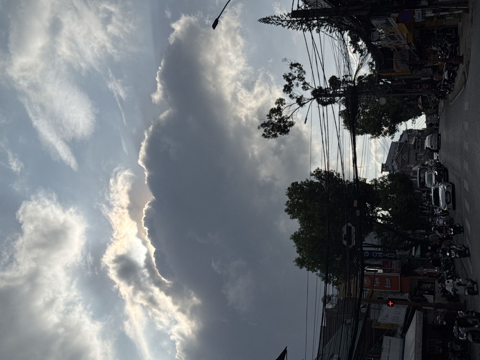 This image, taken in Phường Xuân Hương, Đà Lạt, Vietnam, depicts an urban street scene under a dramatic sky. The sky dominates the left side of the frame, featuring a large, dark cloud with its edges brightly illuminated, indicating the sun is either behind it or breaking through. Other parts of the sky show lighter, wispy clouds against a pale blue.
The right side of the image reveals a busy streetscape. A dense network of overhead power lines and communication cables crisscrosses the upper and middle portions, creating an intricate web. Silhouetted trees with dark foliage are visible, interspersed among the wires and utility poles. Below, the street is lined with numerous parked motorbikes and several cars. Buildings with varying designs are visible along the road, some partially obscured by the urban infrastructure and trees.
Notable details include a red traffic light on the right. Several signs with Vietnamese text are visible: a prominent red sign reads "Tiệm Bánh" (Bakery shop), a smaller sign displays "24h," a blue sign indicates "100m," and another blue sign shows "G CHAU" partially. The lighting suggests it could be late afternoon or early evening, or a heavily overcast day with the sun momentarily piercing through the clouds. The scene captures a typical, vibrant urban environment with a significant presence of motorbikes and overhead wiring characteristic of many Vietnamese cities. No specific human activity is in progress, but the parked vehicles imply a bustling area.