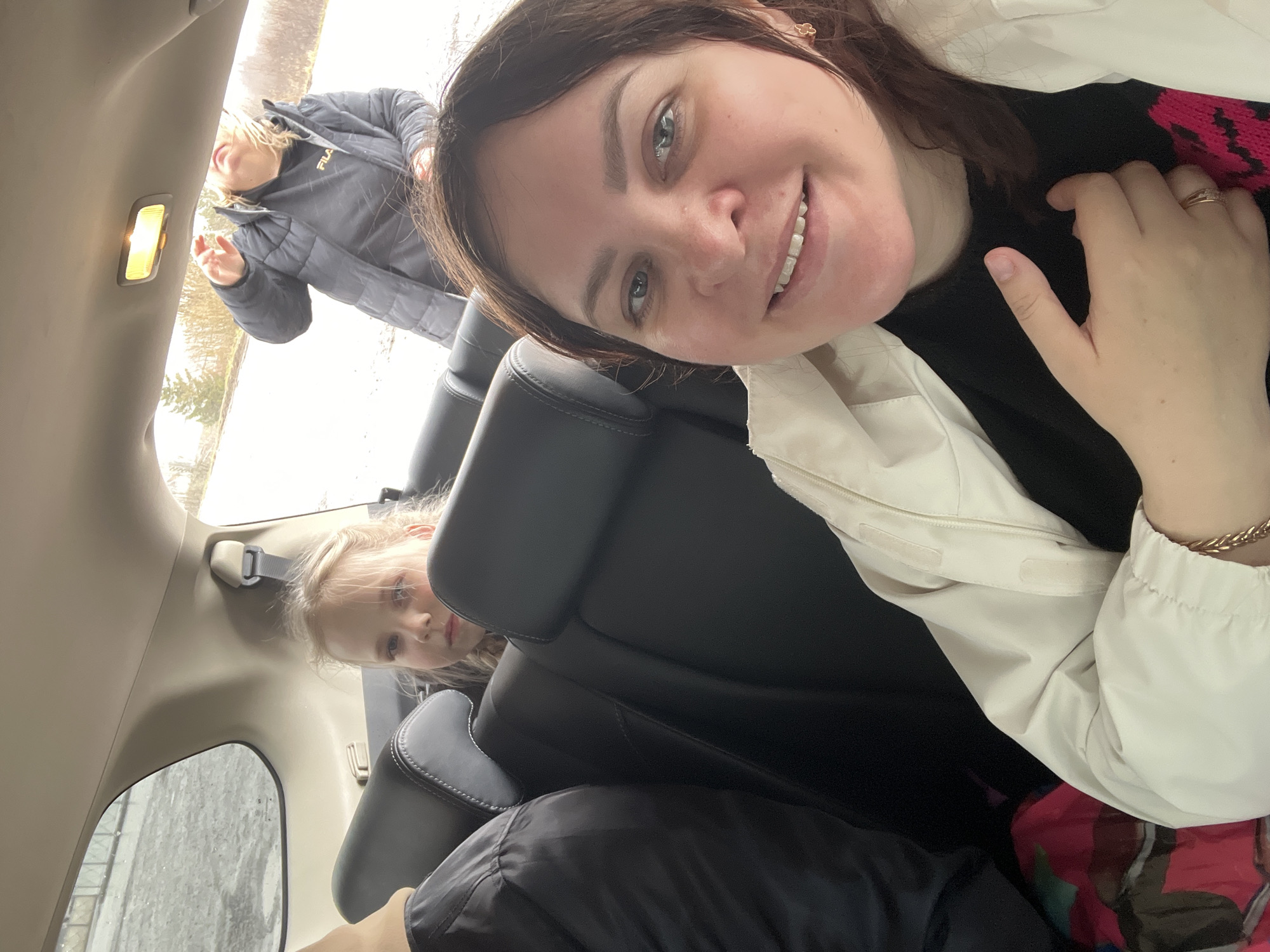 The image captures a selfie taken inside a vehicle. A woman with dark hair and a white jacket is smiling at the camera, holding her hand up near her face. Behind her, a younger blonde girl is peeking between the seats. Further back, near the open trunk of the vehicle, another person in a grey jacket is partially visible. The background suggests an outdoor environment with a bright sky. The location is Kosoturikha, Russia.