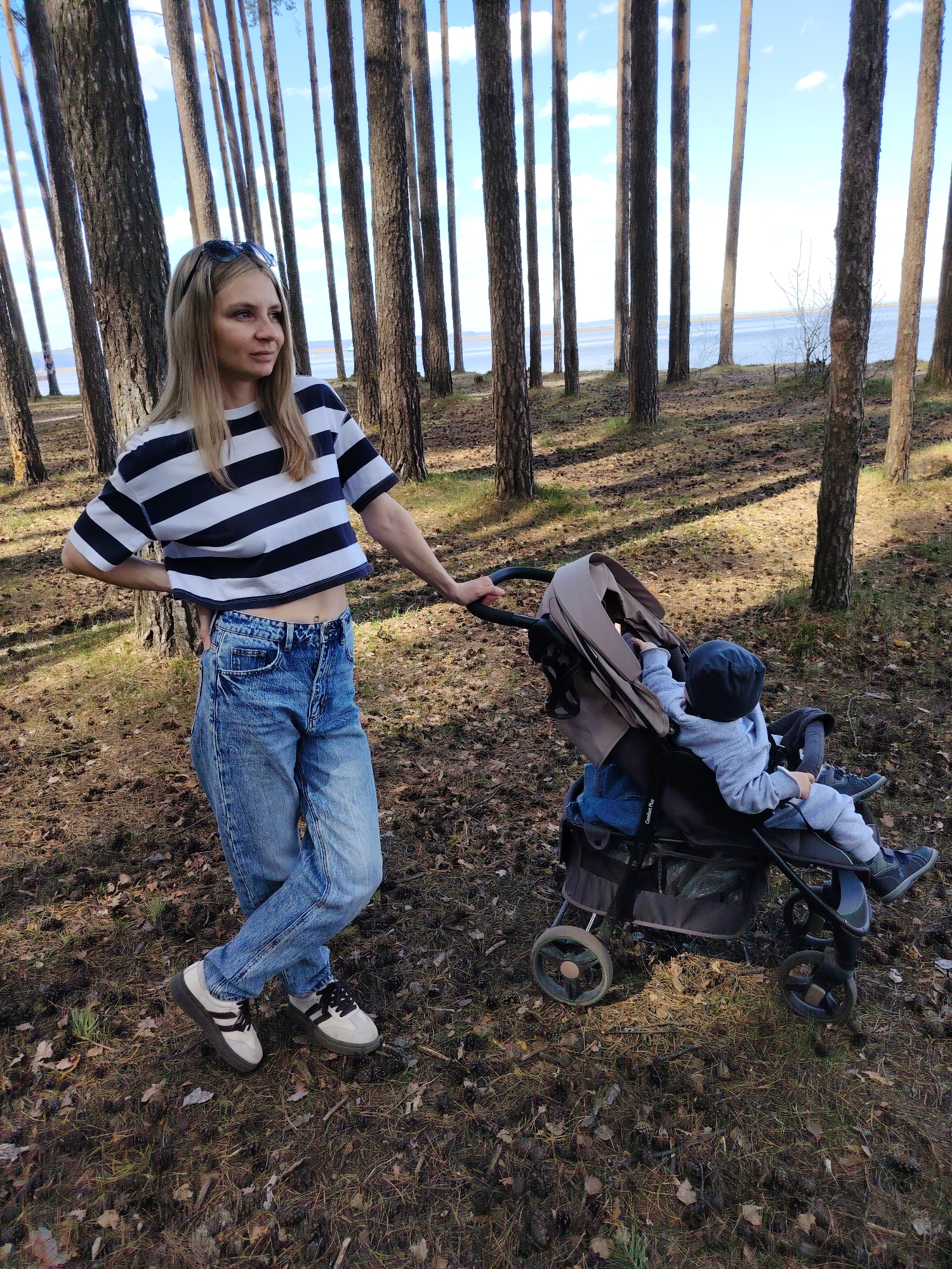 A woman stands next to a baby stroller in a pine forest. The woman has long blond hair and sunglasses on top of her head. She wears a navy and white striped crop top, blue jeans, and platform sneakers. She rests her right hand on her hip and her left hand on the stroller. A baby in a gray hoodie and dark beanie sits in the stroller. The forest floor is covered in pine needles. Trees line the background, and water is visible in the distance. The location is likely near Chaikovsky, Russia, based on the prevalence of pine forests in the region.