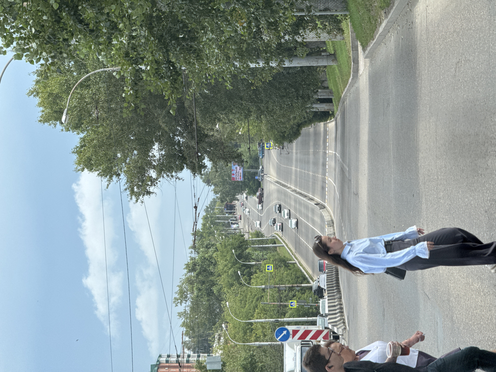 This is a medium-angle, eye-level shot of a multi-lane road in Irkutsk, Russia, with a slight downward slope. The road is paved and has white dashed lines marking the lanes. A pedestrian crossing is visible in the foreground, with three people crossing. Two individuals, a man with glasses and a woman with short hair, are walking from left to right, with the woman holding a clear plastic cup. A third person, a woman with long dark hair, wearing a light blue shirt and dark pants, is also walking from left to right, further ahead of the other two.
On the right side of the road, a line of mature trees with dense green foliage borders the sidewalk and a grassy embankment. The trees are spaced out and appear healthy. Beyond the trees, a light pole with a street lamp is visible. Several other street lamps are spaced along the road, some curving over the lanes. On the left side of the road, there is a concrete barrier with black and white stripes separating the lanes from a grassy area. Further in the distance, on the left, there is a building with a red brick facade.
The road is moderately busy, with several cars and a red pickup truck visible in various lanes, traveling in both directions. Traffic lights and directional signs are also present along the road. The sky is partly cloudy, with patches of blue and white clouds. Overhead, power lines are strung across the scene, supported by poles. A billboard with Russian text, including the word "БОЛЬШЕ" (meaning "more"), is visible in the distance on the left side of the road. The overall environment suggests an urban setting on a clear day.