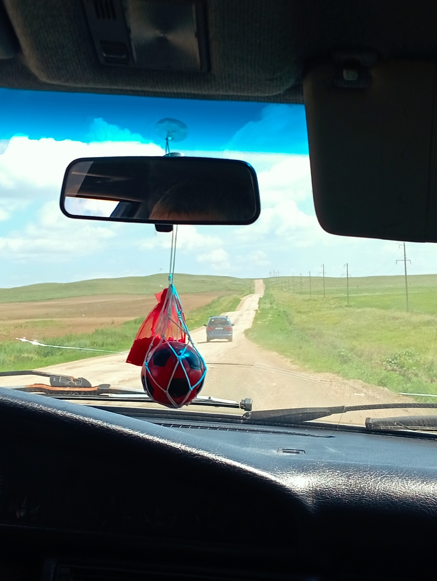The image depicts a view from inside a vehicle driving on a dirt road. A rearview mirror hangs from the windshield, reflecting the sky and a person's head. A red and black soccer ball, attached to a string, dangles in front of the mirror, with a red cloth also attached. The road stretches into the distance, lined with green fields and utility poles. Two vehicles are visible on the road; one in the distance and another closer to the foreground. The sky is blue with scattered clouds. The overall scene suggests a rural or steppe landscape. The location is identified as Aleksandrovka, Kazakhstan.