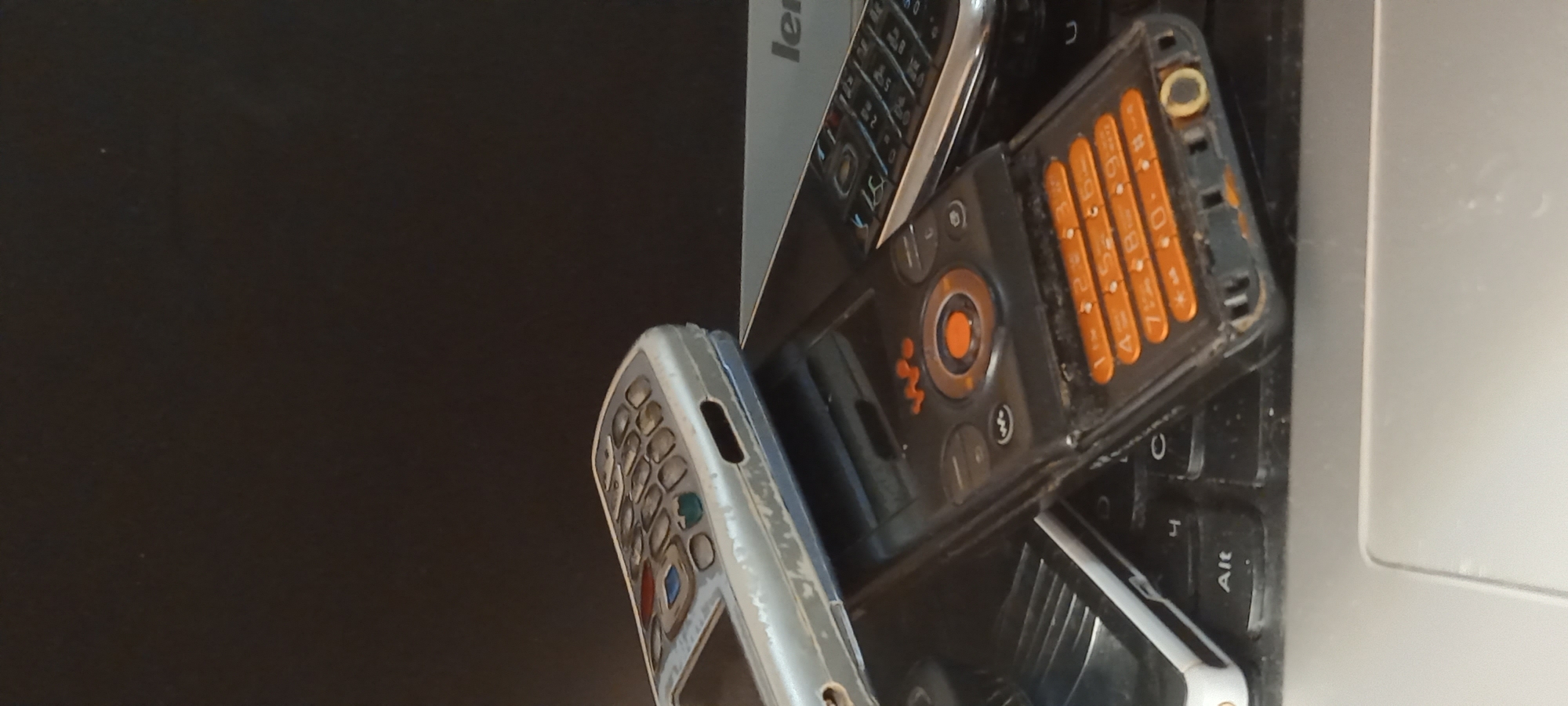 The image captures a collection of several old mobile phones placed on a laptop keyboard, likely on a dark surface in Kryvyi Rih, Ukraine. The scene showcases vintage electronic devices, displaying signs of age and use.
In the center, a black mobile phone is partially open, revealing an orange numeric keypad with white numbers from 1 to 9 and 0, along with associated letters. This phone features a distinctive circular navigation pad with a bright orange central button and a visible "W" logo, suggesting it is a Sony Ericsson Walkman series device. The phone's plastic casing appears worn, with visible dust and scratches.
To the left, a silver-colored mobile phone, also showing considerable wear, displays its numeric keypad. Its surface is scuffed and faded. A third, darker mobile phone is partially visible underneath the black phone.
These devices rest on the black keyboard of a laptop, where keys such as 'C', '0', '4', and 'Alt' are discernible. The "Lenovo" brand name is visible on the edge of the laptop's screen or lid. The background is a plain dark surface, contrasting with the items in the foreground. The lighting suggests an indoor setting, likely during the day. No people are present, and no specific activity is taking place; the image focuses solely on the arrangement of these older electronic gadgets.