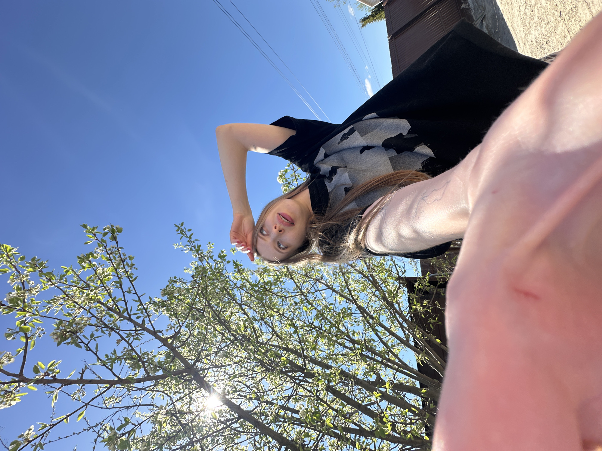 A photograph taken from a low angle shows a woman with light skin, light brown hair, and visible eyelashes, wearing a patterned dark shirt. Her arm is raised, obscuring the lower right of the frame. In the background, there is a clear blue sky and a tree with white blossoms. There is also a building, and power lines are visible in the upper left. The photograph was taken in Chaikovsky, Russia.