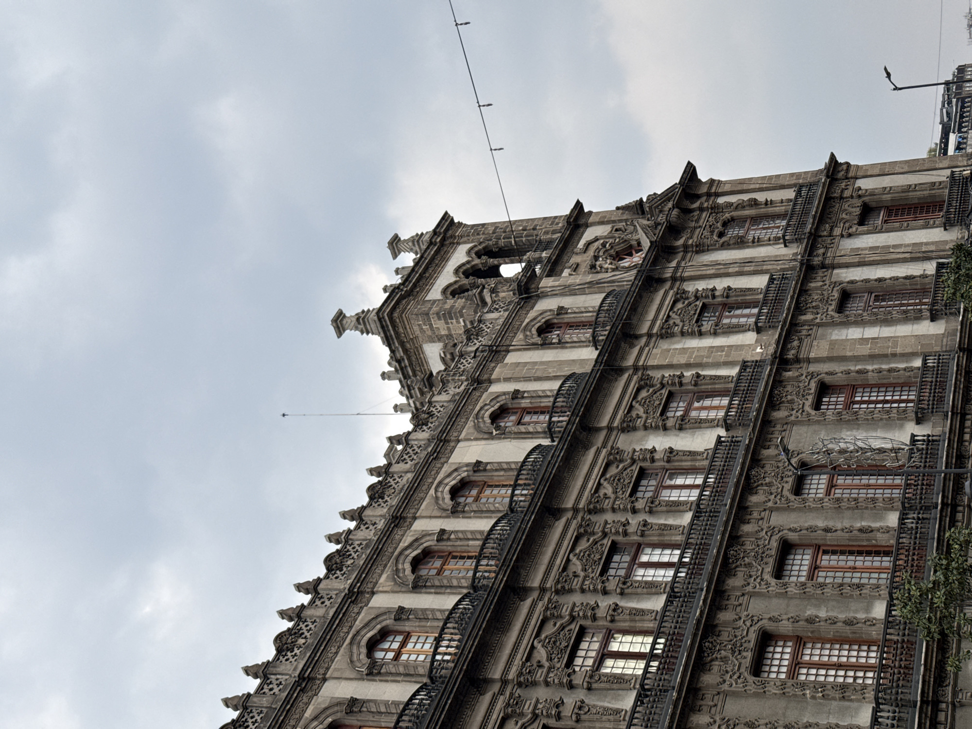 The image captures a low-angle view of a building facade in Mexico City, Mexico, under a cloudy sky. The building displays ornate stonework and architectural details, including rows of windows with dark frames and iron balconies. A metal support and electrical cable cut across the upper portion of the image.
