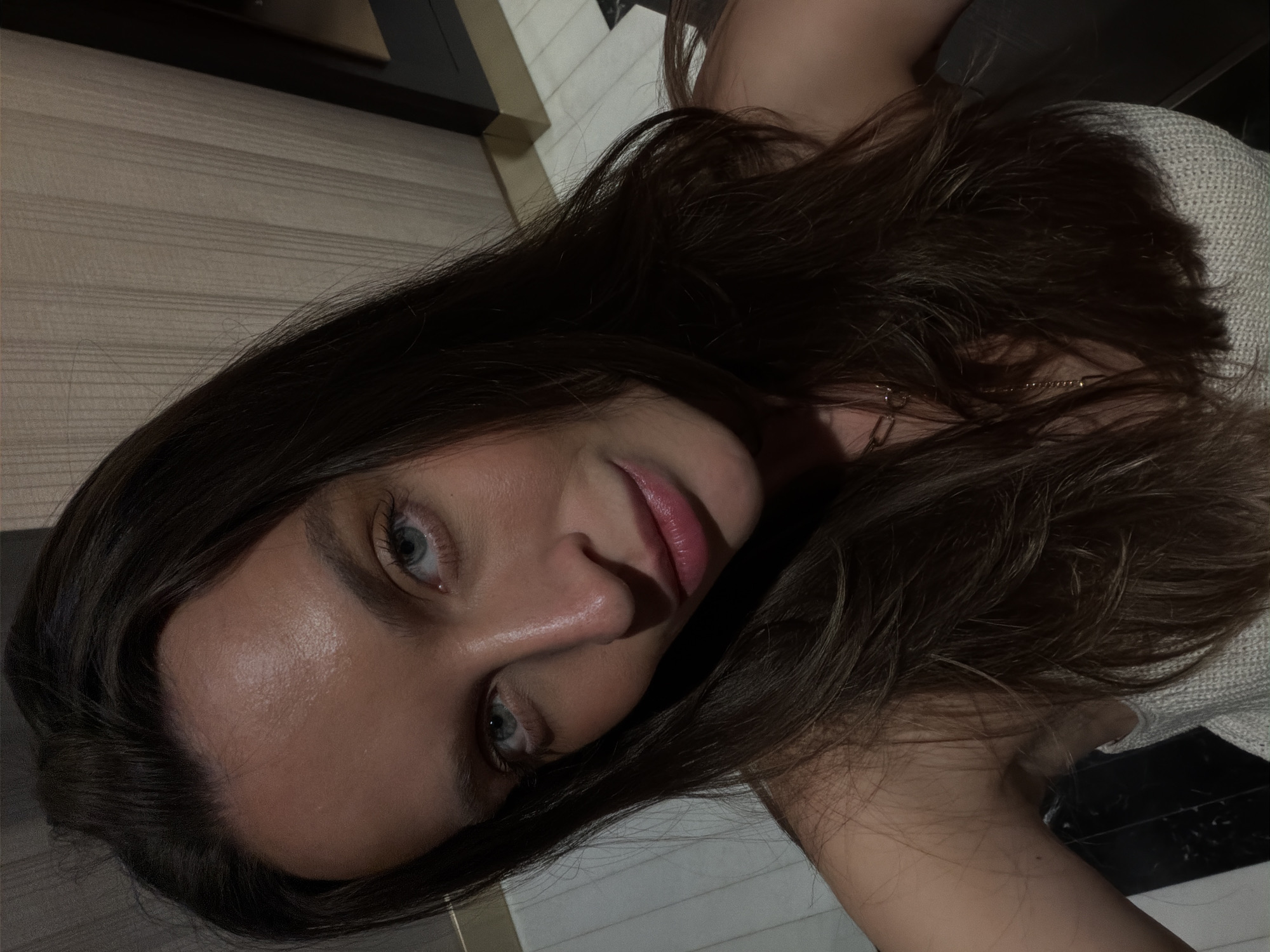 A young woman with long, brown hair is looking directly at the camera. Her eyes are light blue. She has fair skin, and her lips are pink. She is wearing a light-colored top. The background includes a wall with horizontal wood-like panels and a dark, shiny surface. The location is not discernible from the image.
