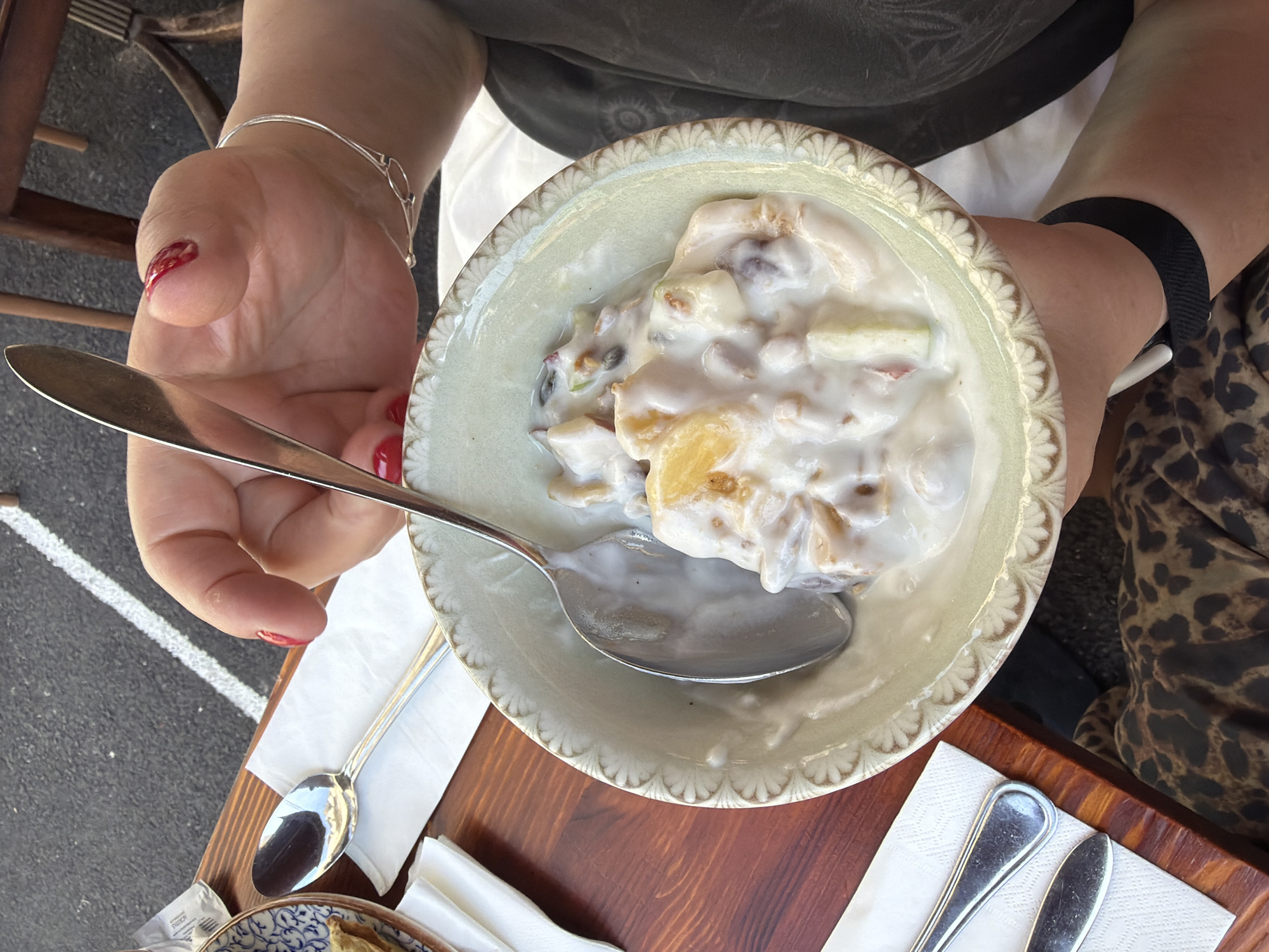 The photograph shows an overhead view of a person holding a bowl of fruit salad with a spoon in it. The bowl is light green and decorated with a scalloped edge. The fruit salad appears to be in a white, creamy sauce. The person's left hand is holding the bowl from the left side and holds the spoon. The person is wearing a silver bracelet on their left wrist and a black fitness tracker on their right wrist. The person is sitting at a wooden table. Next to the bowl, on the table, are some white napkins and other silverware. The backdrop includes the person's leopard-print pants and a glimpse of the street with a white line marking. The location appears to be outdoors and may be in Tel Aviv, Israel due to the presence of a possible Israeli breakfast or brunch item.