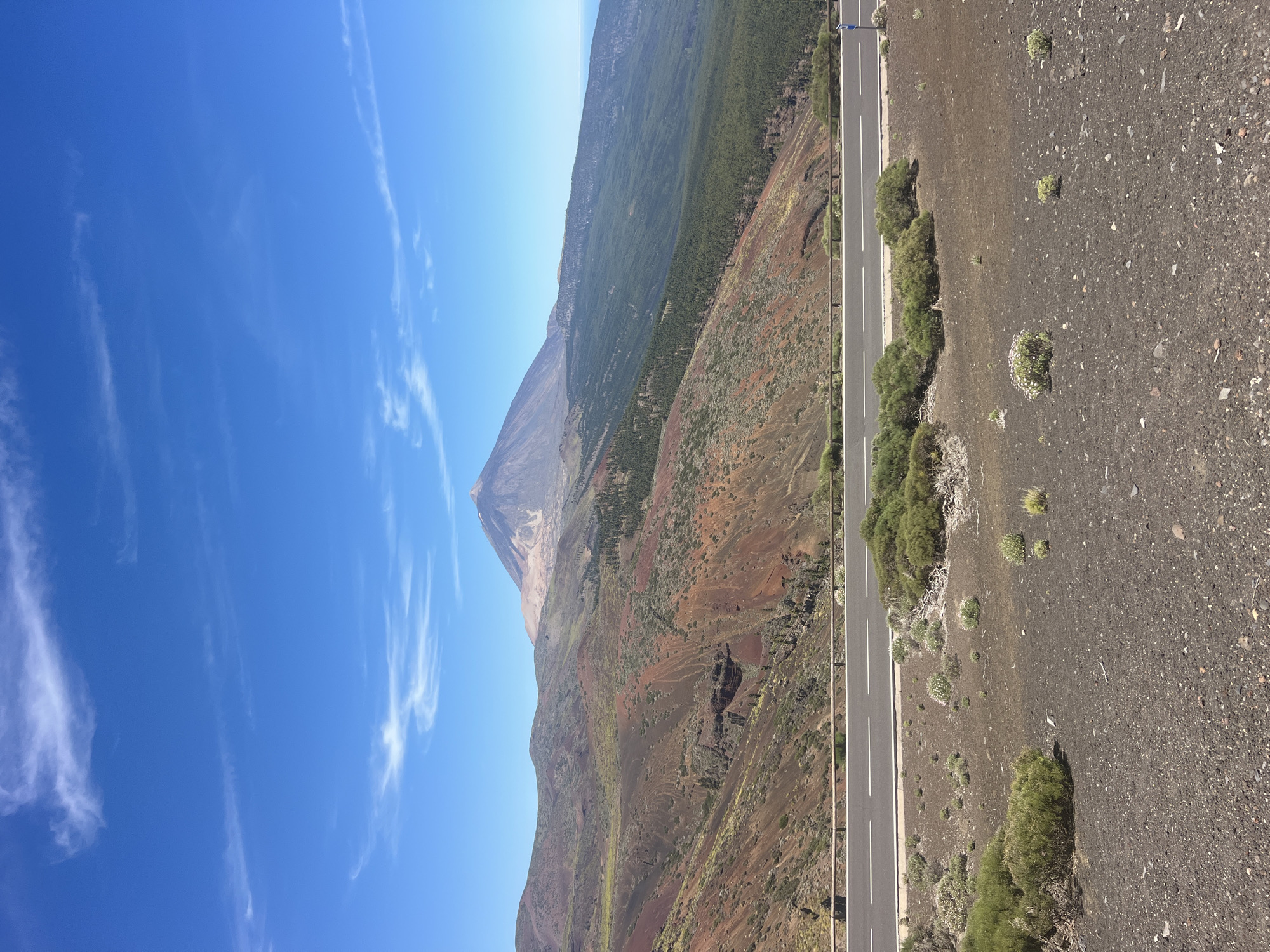 This media file captures a sweeping, high-angle view of a volcanic landscape under a clear, bright blue sky with wispy clouds. The scene is dominated by a conical mountain peak, likely a volcano, with a distinct summit that appears snow-capped or bathed in light. The mountain's slopes display a range of colors, from barren reddish-brown volcanic soil to areas covered in dense green vegetation, possibly pine trees, creating a stark contrast.
A paved road with white lane markings runs diagonally through the frame, bordered by a metal guardrail. The road is flanked by sparse, hardy vegetation growing out of the dark, rocky terrain. Small, low-lying green plants and bushes are scattered across the foreground, suggesting a dry, arid environment. The ground is covered in dark volcanic gravel and pebbles. In the distance, more rolling hills and mountainous terrain can be seen, covered in a mixture of vegetation and bare rock.
The image appears to have been taken during the daytime, judging by the strong sunlight and the clear sky. The lighting suggests it could be mid-morning or early afternoon. There are no visible people in the frame. The overall impression is one of natural grandeur and rugged beauty, highlighting the unique volcanic topography of the region, possibly Tenerife in La Orotava, Spain, given the location context. The image conveys a sense of scale and isolation, with the vastness of the landscape and the imposing presence of the volcano. No text is visible in the image.
