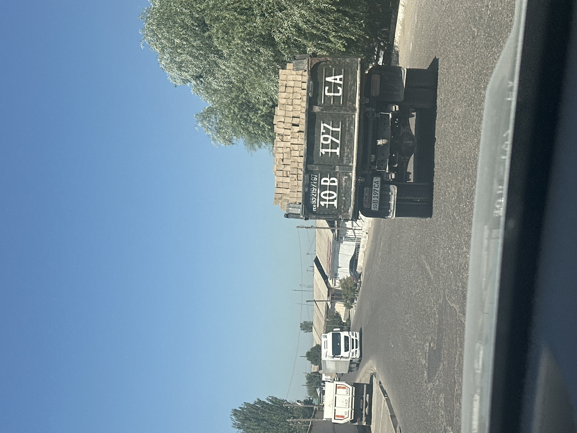 The image depicts a street scene in Yallama, Uzbekistan. The primary focus is on a dark-colored truck in the foreground, loaded with what appears to be bricks. The truck has a license plate that reads "10 B 197 CA". To the left of this truck, a white truck is visible, as well as another white truck further down the road. The street is paved and appears to be in a commercial or industrial area, with buildings lining the right side of the road. Trees and vegetation are present along the sides of the street and in the background. The sky is clear and blue, indicating a sunny day. The perspective suggests the photo was taken from another vehicle traveling on the same road.