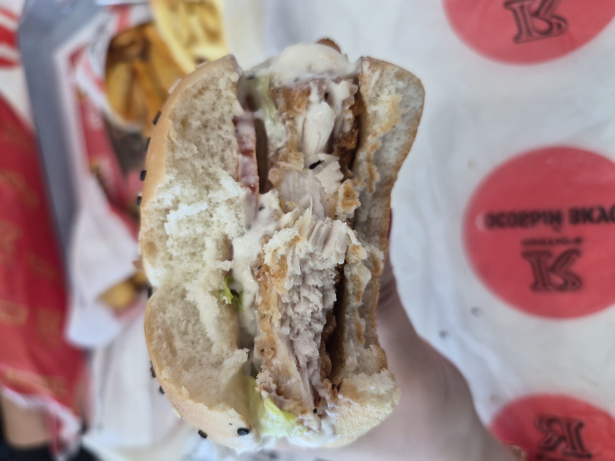 The image shows a partially eaten chicken sandwich. The sandwich is cut in half and held in someone's hand, revealing layers of fried chicken, lettuce, tomato, and mayonnaise. The bun has black sesame seeds on its surface. The background includes a container of french fries, and a white takeout bag with red circular logos bearing a stylized "K" and text in Russian, likely "Скорпион БКАС", suggesting the restaurant is named "Scorpion BKAC". The takeout bag suggests the sandwich was likely purchased in Tobolsk, Russia.