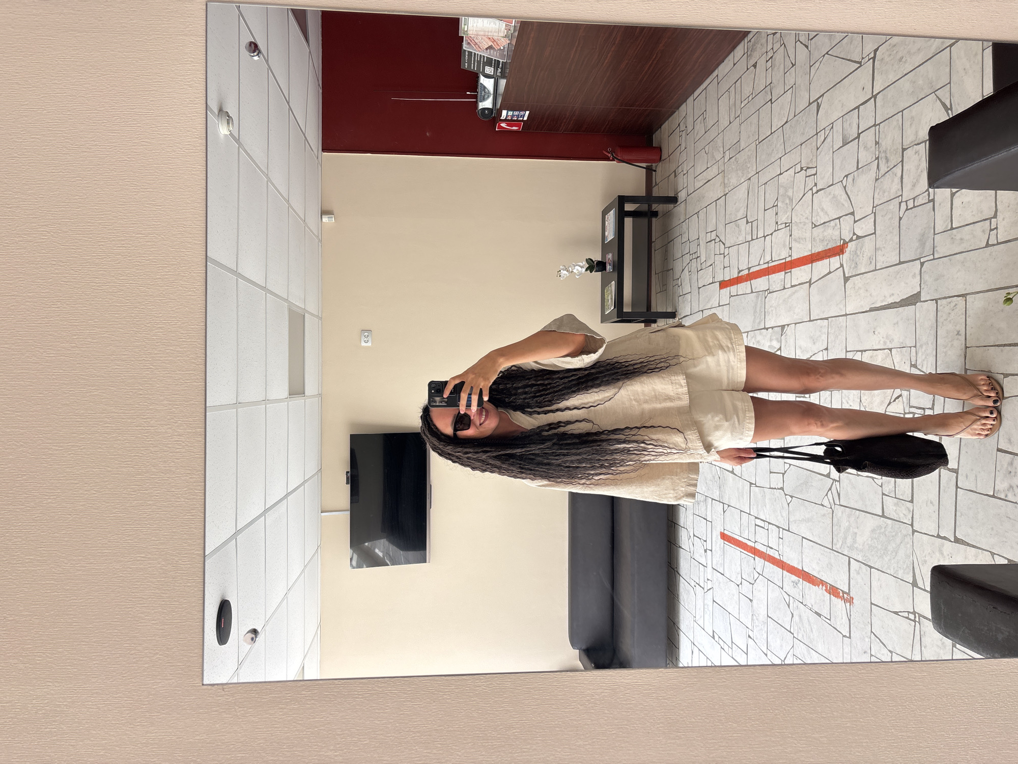 The image is a mirror selfie taken by a person with long, dark, braided hair (dreadlocks or box braids). The person, who appears to be a woman, is wearing sunglasses and a light-colored (possibly linen) short-sleeved top and matching shorts, indicative of a casual, warm-weather outfit. She is holding a black smartphone to take the picture, and her mouth is visible, suggesting a smile. Her feet are visible, wearing light-colored flip-flops, with dark nail polish on her toenails. She is also holding a black bag in her left hand.
The setting appears to be an indoor commercial or public space, possibly a waiting area or salon, reflected in the large mirror. The floor is covered with light-colored, irregularly shaped tiles, creating a mosaic pattern. A prominent orange-red line is painted across the floor, which might indicate a social distancing marker or a designated boundary. The walls are painted in two tones: a light beige and a darker red. Visible in the reflection are a black, wall-mounted flat-screen television, a dark-colored sofa or bench, a small black table with a white artificial flower arrangement, and an electrical outlet. Above the table, on the red wall, there are some papers or signs with text that is too small to decipher, and a red fire extinguisher is mounted below them. The ceiling is made of white acoustic tiles, and two light fixtures or sensors are visible there. The mirror itself is bordered by a light beige textured wall. The general ambiance is well-lit by interior lighting. No specific time of day is indicated, and the location is Chaikovsky, Russia.