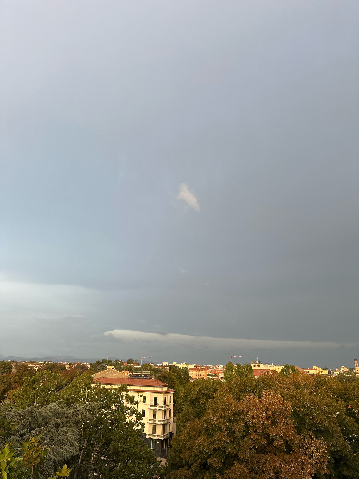 A high-angle shot captures an urban landscape in Parma, Italy, under a moody, overcast sky. The foreground is dominated by the lush, green foliage of trees, with some exhibiting the warm hues of autumn. A prominent, light-colored building with a terracotta roof stands out amidst the greenery, featuring multiple stories with visible windows. In the background, a cityscape unfolds with numerous buildings, some with red tile roofs, interspersed with more trees. The sky transitions from a lighter gray near the horizon to a darker, more ominous gray further up, with a single, small, white cloud visible in the upper portion of the frame. Faint outlines of distant hills can be seen on the horizon.