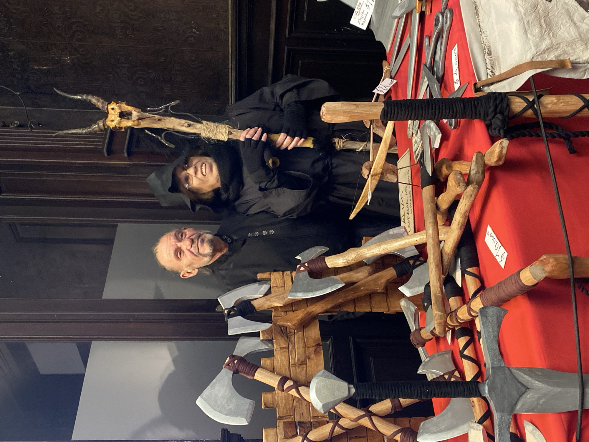 The image shows two people standing behind a display of hand-crafted weapons, potentially at a fair or market. The man on the left, with a shaved head and wearing a black tunic, stands slightly behind the woman. The woman on the right wears a black hooded robe and holds a staff topped with animal horns. In front of them are various handcrafted weapons including axes, swords, daggers, and bows, displayed against a red background. The axes are arranged in a layered fashion, some attached to a wooden structure. Price tags are visible on some items. The setting appears to be indoors, possibly a historic building with dark wooden paneling in the background. The precise location in the city of Buenos Aires, Argentina cannot be determined from the image.