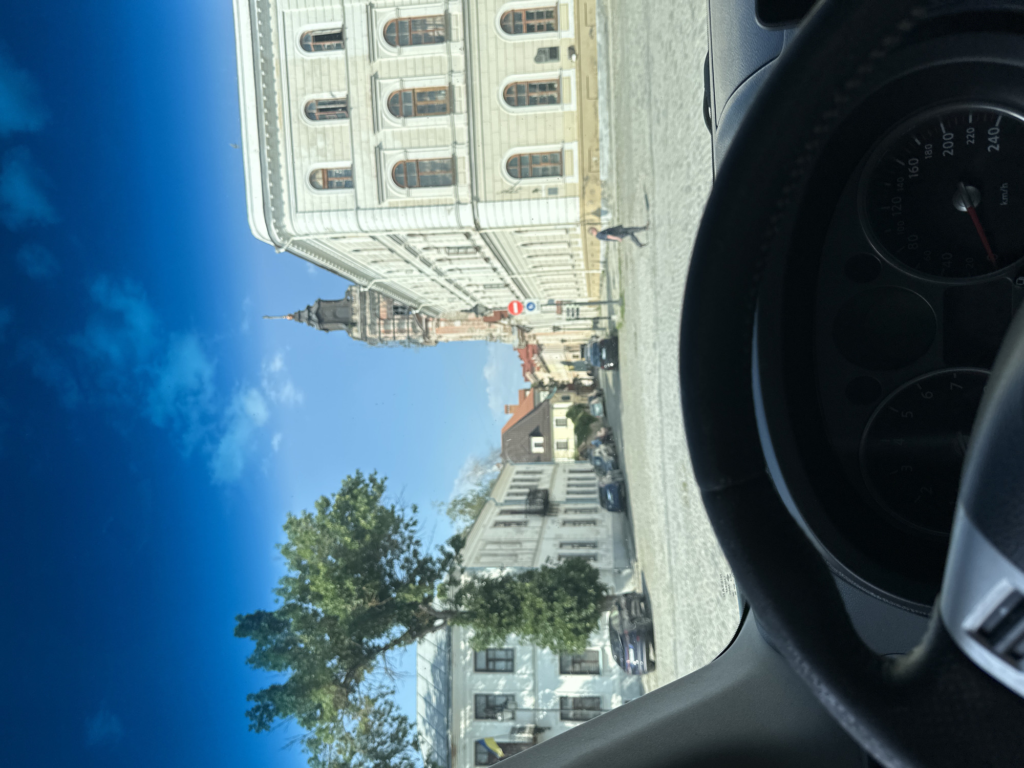 The image is taken from inside a car, looking out at a European city square. The foreground shows the steering wheel and a portion of the dashboard with a speedometer. The street is paved with cobblestones.
On the left, there is a white building with multiple windows and a balcony, with a tall green tree in front of it. A Ukrainian flag is visible on the left side of this building. Further down the street, more buildings with traditional architecture are visible, with cars parked along the side.
In the center background, a prominent tower with a dark spire rises above the surrounding buildings. To the right of the tower is a large, ornate building with many windows and classical architectural details, painted in a light color. A person is walking across the square in front of this building. The sky is blue with some white clouds.
The scene is consistent with the city of Kamianets-Podilskyi, Ukraine, known for its historic architecture and cobblestone streets, particularly around the Old Town area.