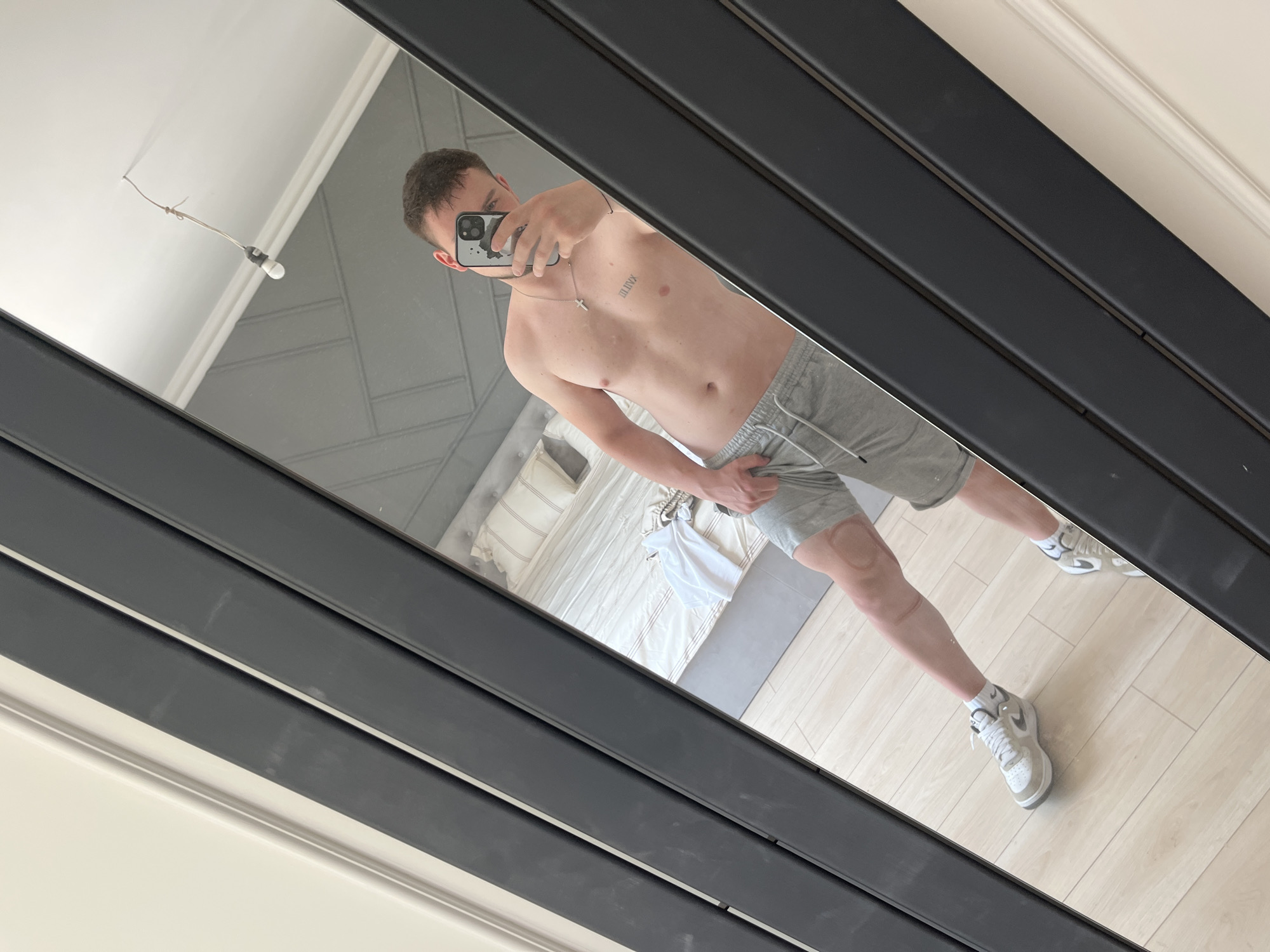 A young man is depicted taking a mirror selfie in what appears to be a bedroom. He is shirtless, wearing grey athletic shorts with a white drawstring, white socks, and light-colored sneakers. He holds a smartphone with a dark-patterned case, which partially obscures his face in the reflection. A silver-colored chain with a small cross pendant hangs around his neck. On his left ribcage, a tattoo of the Roman numerals "M.M.IX" is visible. A faint, circular mark, possibly a cupping therapy mark, is present on his right upper thigh/knee area, alongside a linear scar above it.
The large mirror features a thick, black frame. The reflection reveals a bright room with light-colored wooden or laminate flooring. In the background, a bed with a grey upholstered headboard displaying a subtle geometric pattern is made with light-colored bedding. A white t-shirt or garment rests on the bed. A single bare lightbulb hanging from a wire is reflected in the upper left corner of the mirror. The overall lighting suggests it is daytime. The man is captured in a casual pose for the selfie. The image provides no specific visual cues to identify the location as Войводино, Bulgaria.