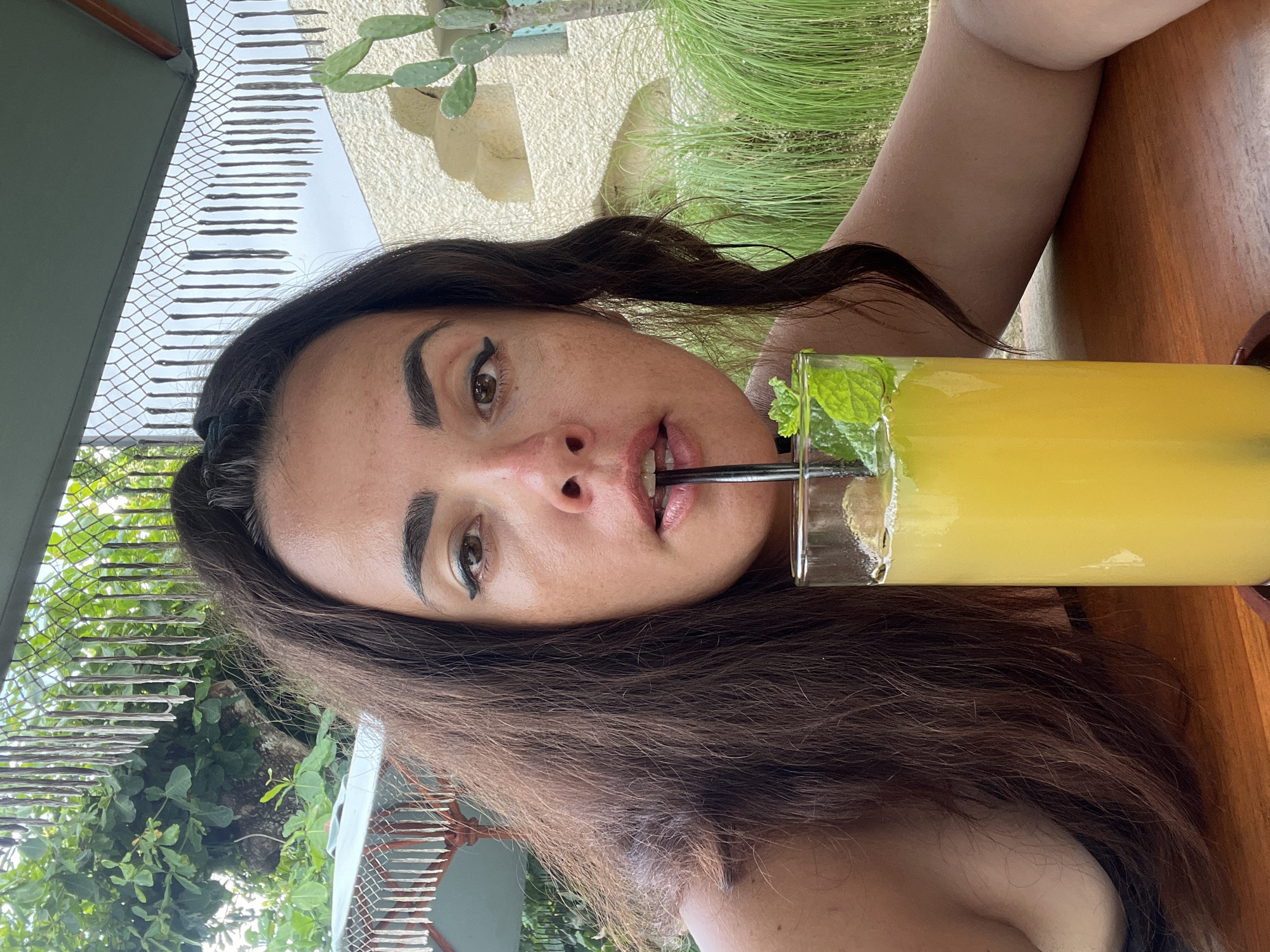 In Kuta Selatan, Indonesia, a woman with long, dark, wavy hair is captured in a close-up selfie. She is seated at a wooden table, leaning forward with her head tilted. Her gaze is directed at the camera, and she is holding a dark straw in her mouth, which is in the process of drinking from a tall glass filled with a yellow beverage and garnished with mint leaves. The woman has distinct winged eyeliner and full eyebrows. Behind her, the environment suggests an outdoor setting with a thatched umbrella overhead, lush green foliage, a textured beige wall, and a prickly pear cactus visible to the right. The overall scene conveys a moment of leisure and refreshment in what appears to be a tropical or resort-like location.