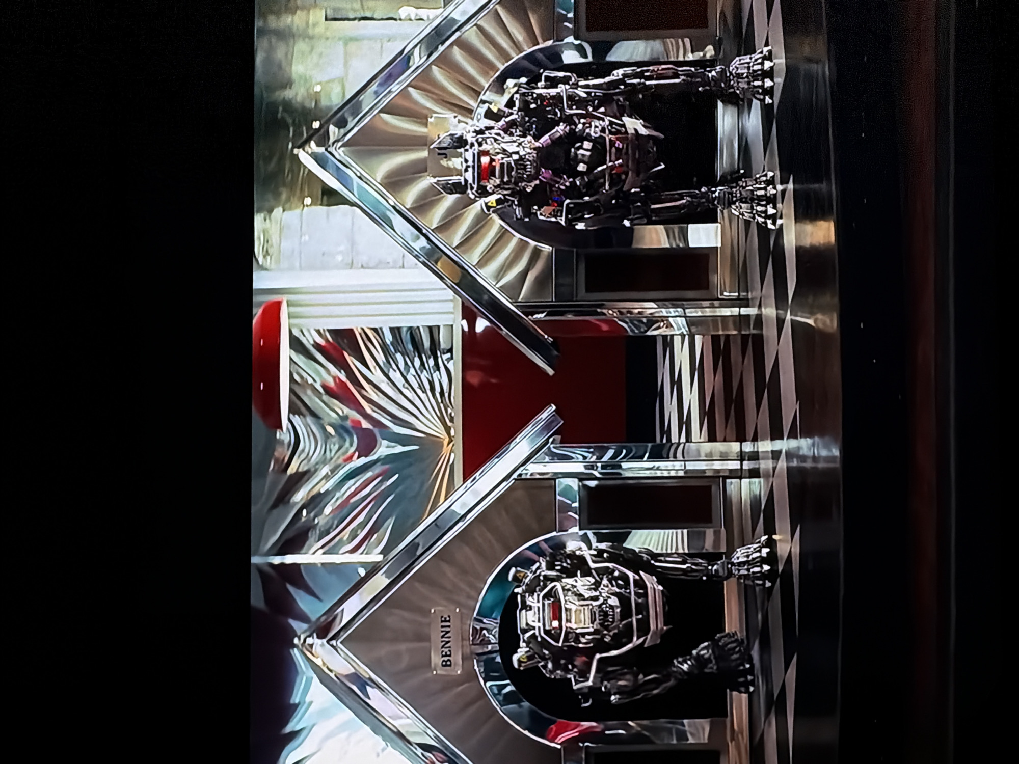 This media file captures an indoor scene featuring two elaborate display cases, each housing a detailed robotic figure, likely in Sofiivska Borshchahivka, Ukraine. The image is oriented vertically, showing the robots standing upright within their enclosures.
The central subjects are two identical or very similar skeletal-like robots, constructed from metallic and mechanical components, resembling sophisticated automatons or sci-fi figures. They appear to be static displays, with intricate internal workings and segmented limbs visible.
Each robot is presented in a large, chrome-plated display case with a distinctive retro or art deco design. The cases feature fan-like, ribbed chrome elements behind the robots, creating a visually striking backdrop. The base and framework of the displays are also polished chrome, reflecting light. The floor beneath and between the displays has a classic black and white checkered pattern, adding to the retro aesthetic.
On the lower display case, a small rectangular nameplate is visible, bearing the text "BENNIE". In the background, hints of a red object, possibly a diner-style booth or chair, can be seen, along with other reflective surfaces and architectural details.
The setting appears to be a commercial space, exhibition hall, or a specialized store, designed to showcase these robotic figures. No human activity or specific event is taking place; the scene is static, focusing on the exhibits. The lighting suggests an artificial, indoor environment.