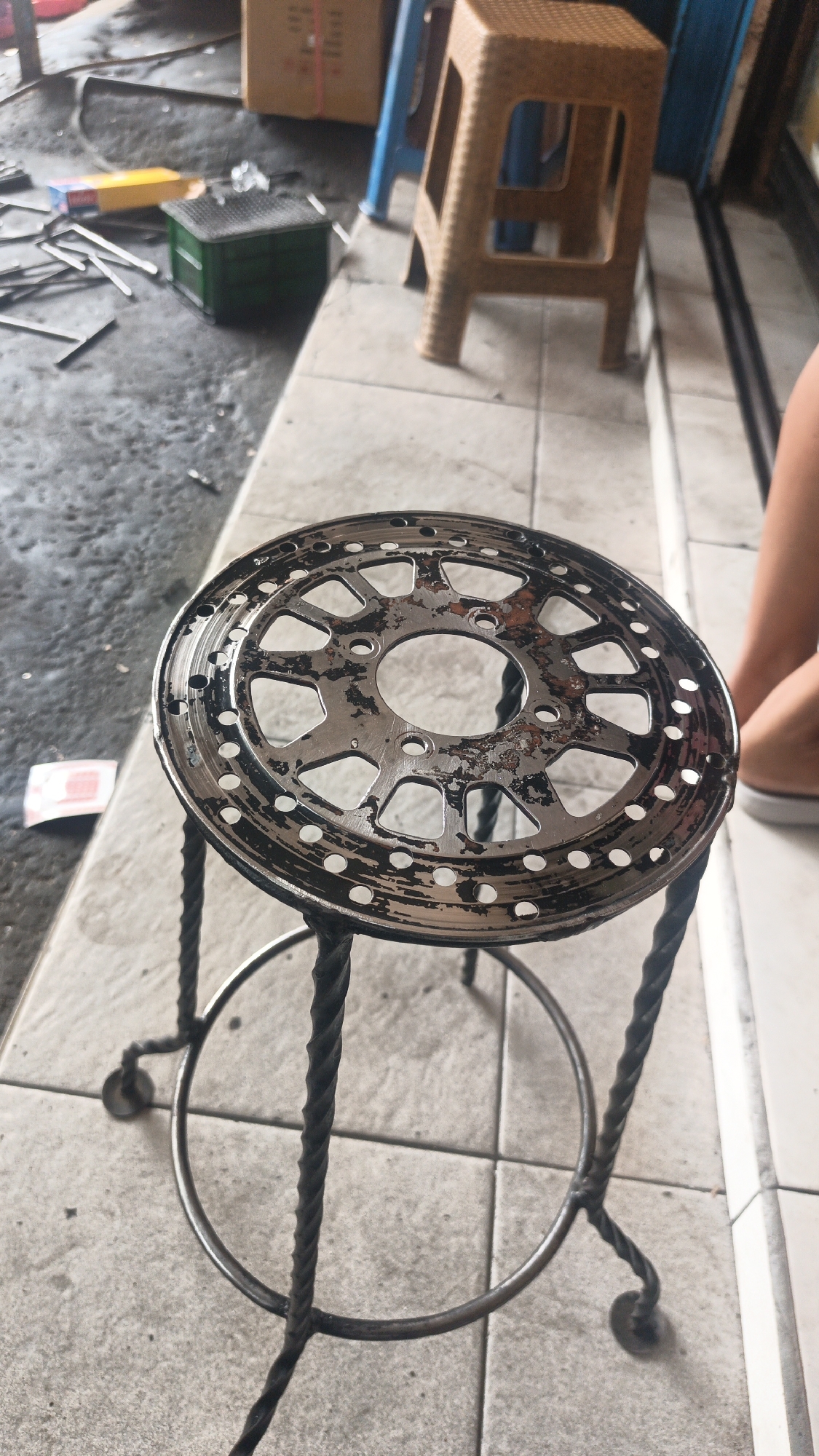 A close-up, slightly angled shot shows a small, round metal stool with a brake disc as its seat. The brake disc is silver with black and rust-colored wear marks and many small holes around its perimeter. The stool's legs are made of twisted metal, forming a circular support near the bottom. The stool sits on a tiled floor. In the blurred background, to the left, are various objects on a rough concrete surface, including metal rods, a yellow and blue box, and a small green wire cage. To the right, a person's legs, wearing shorts and white shoes, are visible, and behind them, part of a brown plastic stool and a blue plastic chair are visible. The scene suggests an outdoor or workshop area in Mlati Kidul, Indonesia.
