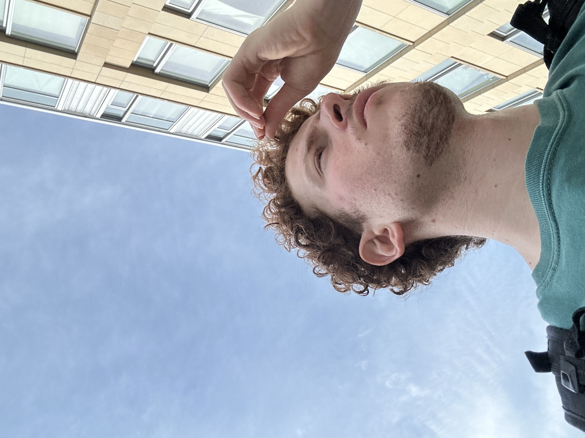 An image, captured from a low-angle perspective, depicts a male individual against a light blue sky with sparse white clouds and a tall, modern building. The subject, a young adult male with light skin, reddish-brown curly hair, and light stubble on his chin and upper lip, is positioned in the lower left portion of the frame, looking upwards and to his right. His right hand is raised, with fingers touching the front of his hair near his forehead. He wears a teal-colored crewneck shirt, and a black backpack strap is visible on his left shoulder.
To the right of the subject and extending upwards, a multi-story building occupies the upper right quadrant. The building features a light beige or tan facade with numerous rectangular windows, mostly clear, reflecting the sky, though some show white interior blinds. The building's architecture suggests a contemporary style, consistent with urban environments. The overall scene is indicative of an outdoor setting during daylight hours within the City of New York, United States of America.
