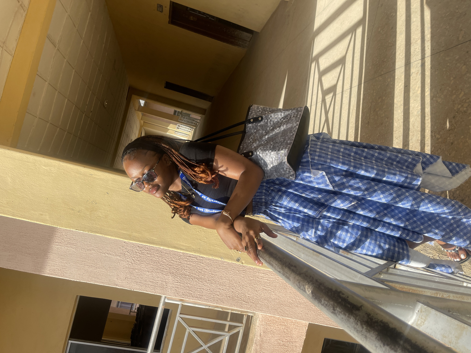 A young Black woman with braided hair and sunglasses is standing on an outdoor walkway, leaning on a metal railing. She is wearing a black crop top and a blue and white plaid skirt. She has a patterned tote bag slung over her shoulder. The walkway appears to be part of a building with yellow and peach-colored walls and a tiled ceiling. The sunlight casts long shadows across the floor, indicating it is daytime. The architecture suggests a possibly institutional setting like a school or university campus, consistent with locations found in Jos, Nigeria, given the presence of the ID card hanging from her neck which says "UNIJOS STUDENTS".