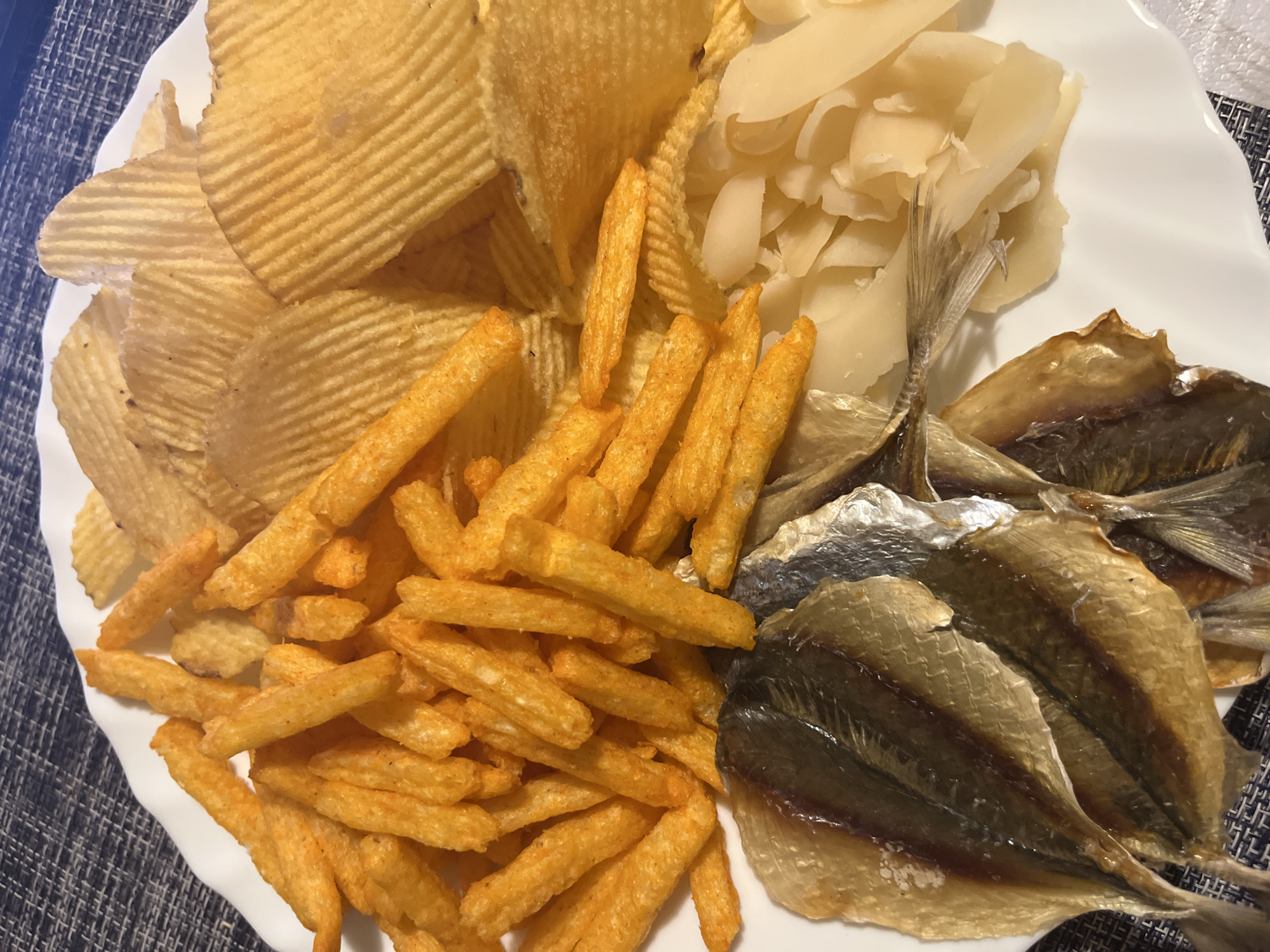 A white plate contains potato chips, orange-colored french fries, sliced bamboo shoots, and small dried fish. The scene is set against a blue patterned surface. No information regarding the location in the city of Раздолье, Russia is ascertainable from the image.