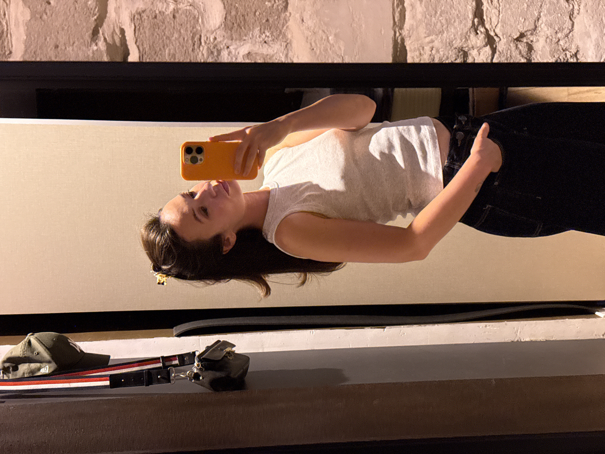 A woman stands in front of a mirror, taking a selfie with her phone. She is wearing a light gray tank top and dark denim jeans. A small black purse and a green baseball cap are hanging on the wall to her left. The background appears to be a room with a stone wall on the right side. The image does not provide any specific details to determine its location in Tel-Aviv, Israel.