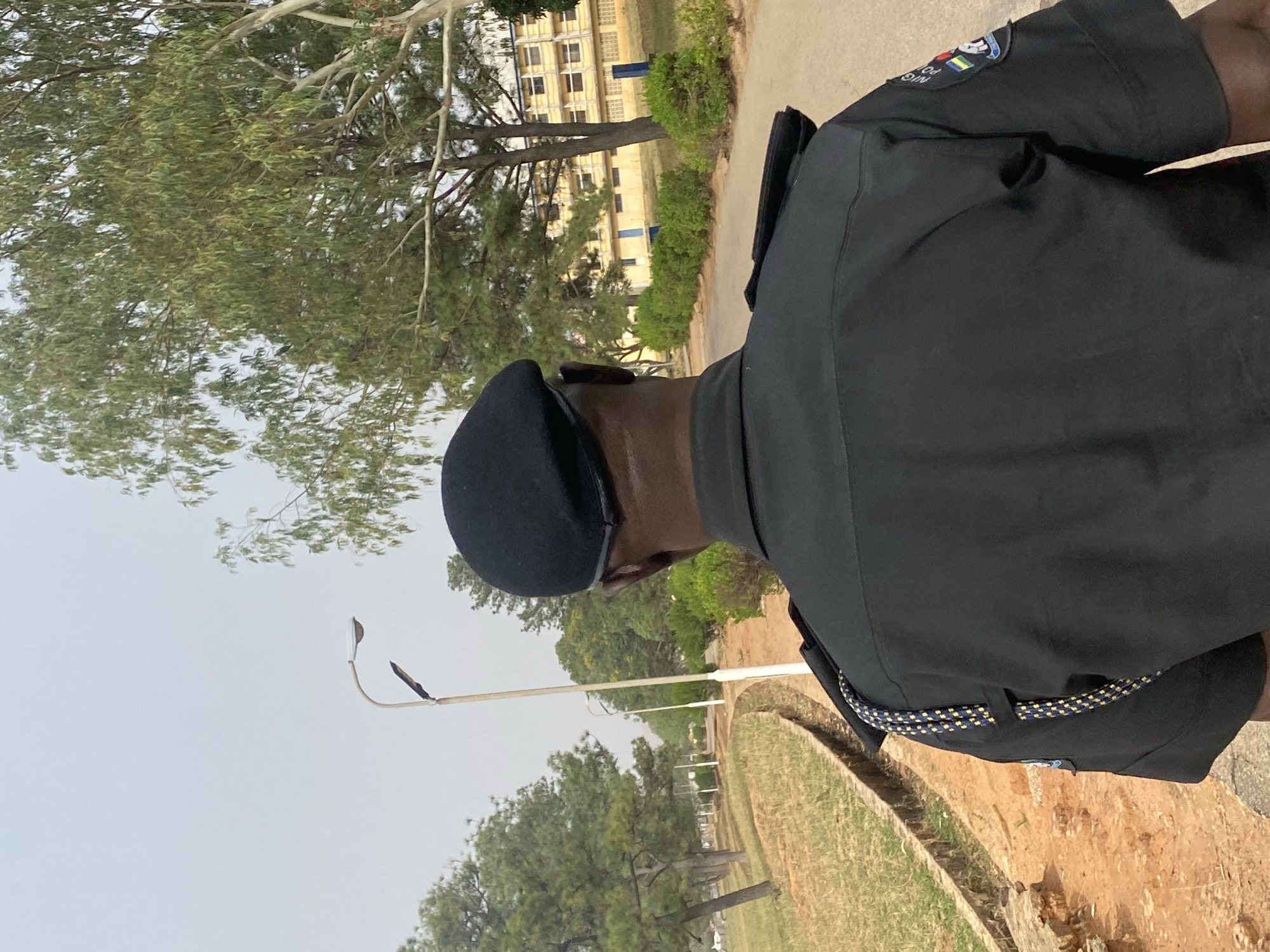 The image captures a person in a police uniform, viewed from the back and slightly to the right, standing outdoors during the daytime. The individual wears a dark beret and a dark, short-sleeved uniform shirt. On the right shoulder of the uniform, a patch is visible, clearly displaying "NIG POL" and "FORCE," indicating the Nigeria Police Force. The patch also incorporates the Nigerian national colors (green, white, green) and an emblem. A patterned belt with yellow and blue/dark dots is worn around the waist.
The background features several light-colored, multi-story buildings with multiple windows, partially obscured by various trees and green bushes. A street light with a curved pole stands in the middle ground on the left. The ground consists of earthy tones, with a concrete or stone-lined drainage channel running along the bottom right. The sky is bright but appears hazy or overcast. The overall scene suggests an institutional or campus environment in Bukuru, Nigeria. The person is not engaged in any specific activity beyond standing or walking through the area.