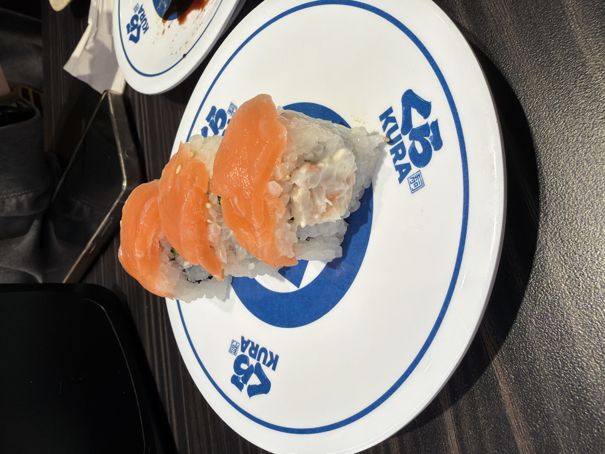 Three pieces of salmon sushi are arranged on a white plate with a blue rim and Kura Sushi branding. The sushi rolls are topped with slices of salmon and have a creamy filling. The plate is placed on a dark, textured surface, possibly a wooden table. In the background, a black object, a smartphone, and a white napkin are visible. Another plate with sushi residue and soy sauce is also present in the upper right corner of the image. The scene appears to be at a Kura Sushi restaurant in Beaverton, Oregon, United States of America.