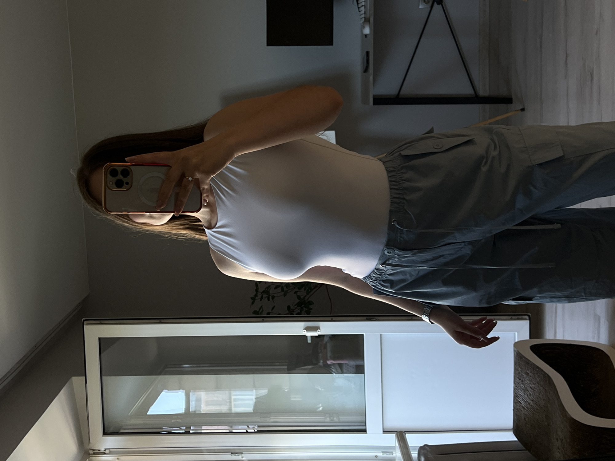 A young woman stands in a room, holding a smartphone to take a mirror selfie. She is wearing a white sleeveless top and grey cargo pants. The room appears to be a domestic interior, possibly an apartment in Ternopil, Ukraine. A large window with white trim is visible to her left, showing a glimpse of the outdoors. To her right, a desk with a computer monitor and keyboard is set up against a grey wall. The overall lighting suggests it is daytime, with natural light entering from the window.