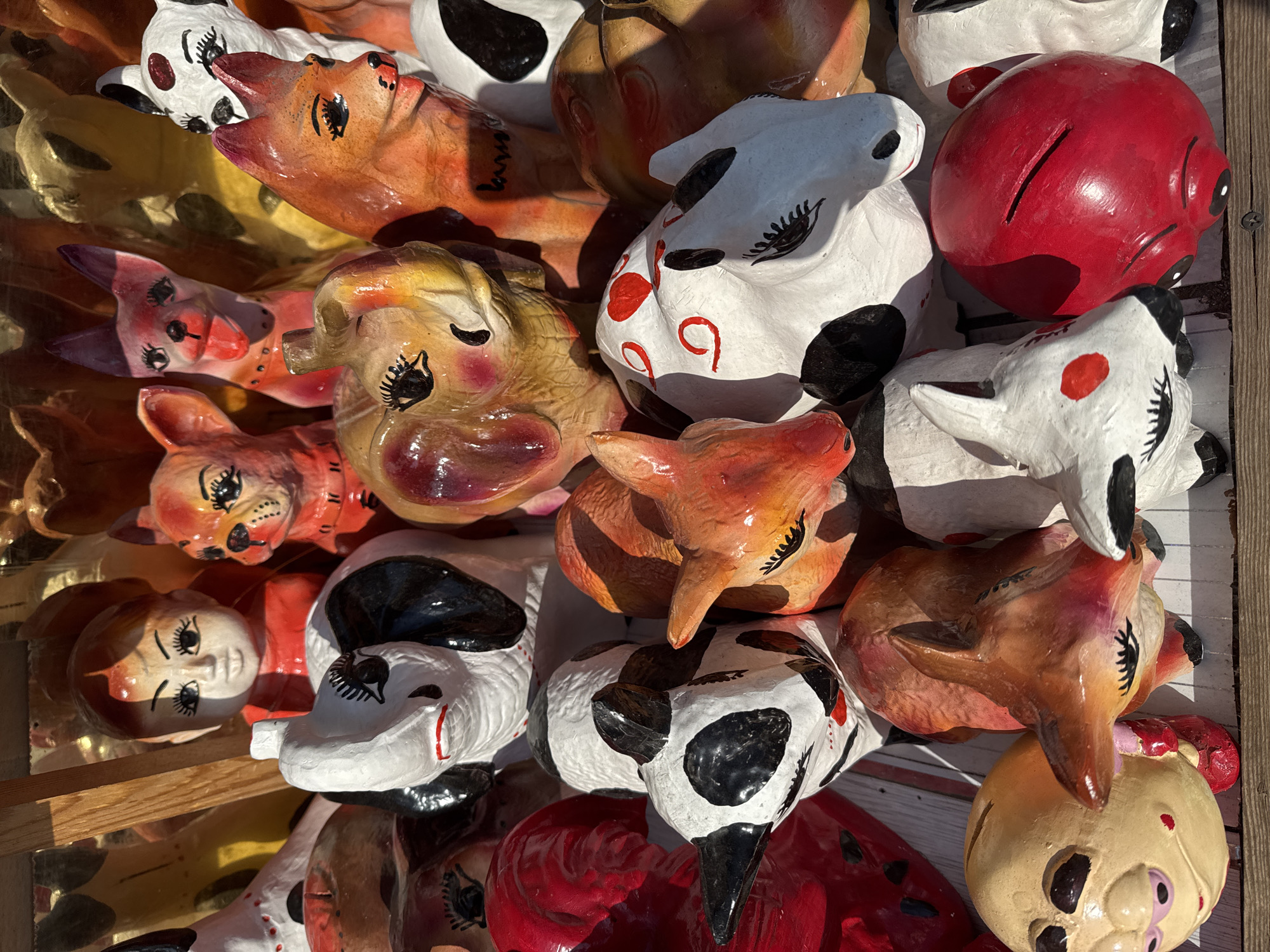 A collection of handcrafted piggy banks, primarily in the shape of animals and a human figure, are displayed outdoors. The objects are made of a material that appears to be clay or papier-mâché, and are brightly painted with various colors and details. The subjects include dogs with exaggerated features, a person with wide eyes and rosy cheeks, and what seem to be elephants and cows. Some of the piggy banks have slots for coins. The display is set against a wooden background, with natural light casting shadows. The location in Baku, Azerbaijan is unknown from the provided image.