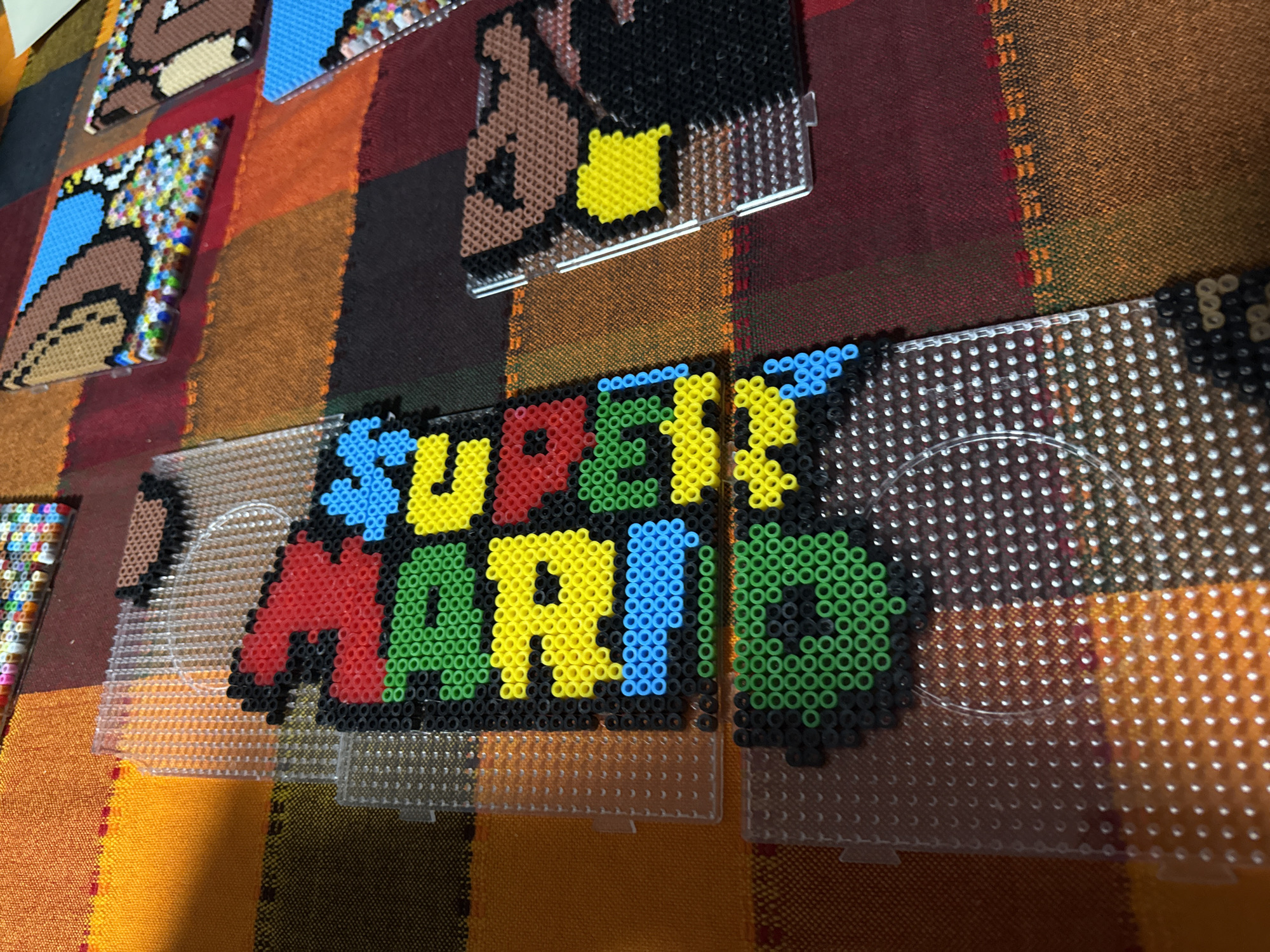 The image shows a partially completed Super Mario logo made of perler beads on a clear plastic pegboard. The "Super Mario" lettering is in various colors, including red, yellow, blue, and green, with black outlines. Several other perler bead art pieces, also on clear pegboards, are visible in the background. The background is a multi-colored striped fabric. The location of the image is presumably in Puebla, Mexico, as indicated by the prompt.