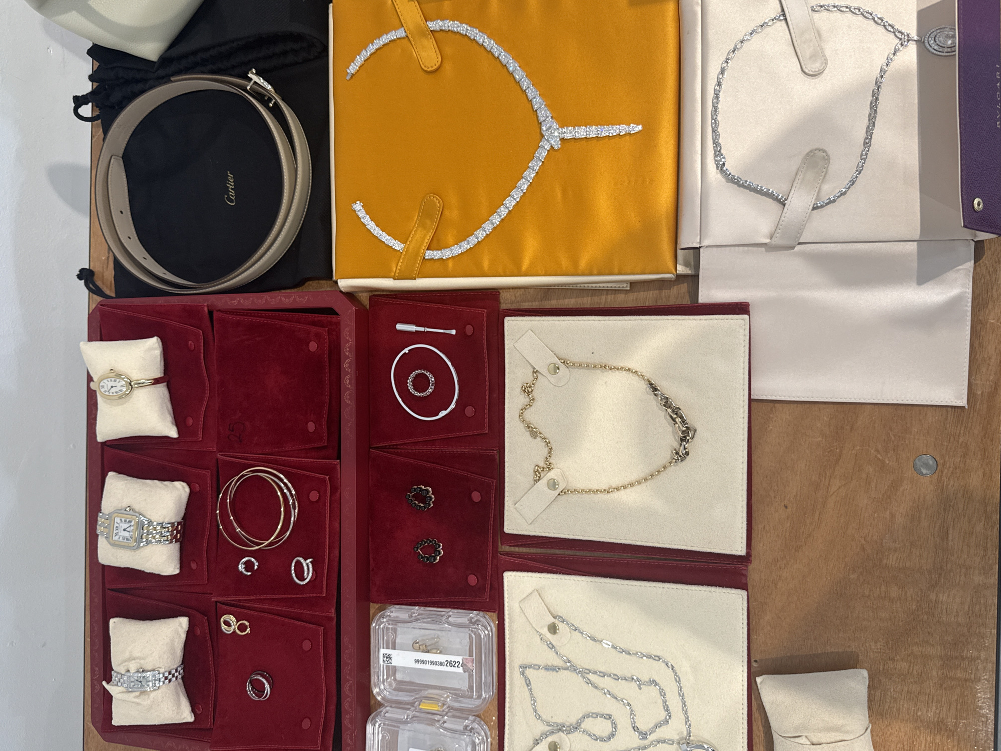 The image depicts a collection of jewelry and accessories displayed on a surface, likely for organization or sale. There are several Cartier items, including a belt, watches, rings, and necklaces, visible by their branding. Other jewelry items include necklaces and rings of varying designs and materials. The items are arranged on different colored backgrounds or in pouches. The location is not specified, and there is no indication that this is in London, United Kingdom.