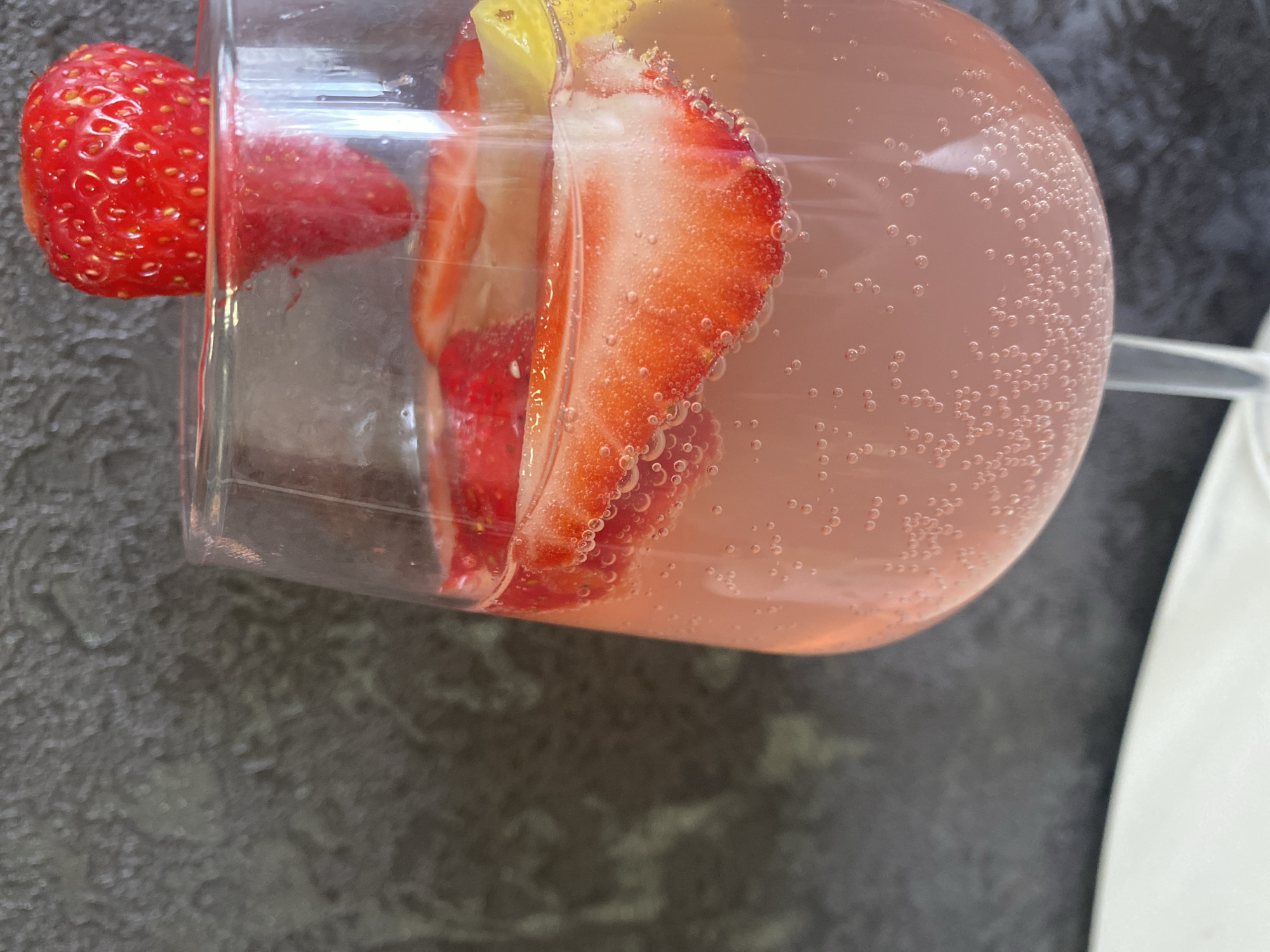 A glass of pink liquid, likely a sparkling beverage, is filled with bubbles and garnished with fruit. A whole strawberry is positioned on the edge of the glass. Inside the glass, there are slices of strawberries and a piece of yellow citrus, possibly a lemon. The glass sits on a dark, textured surface. A portion of a white object, possibly a plate or cloth, is visible on the right side of the image. There is no indication that the location is Izhevsk, Russia.