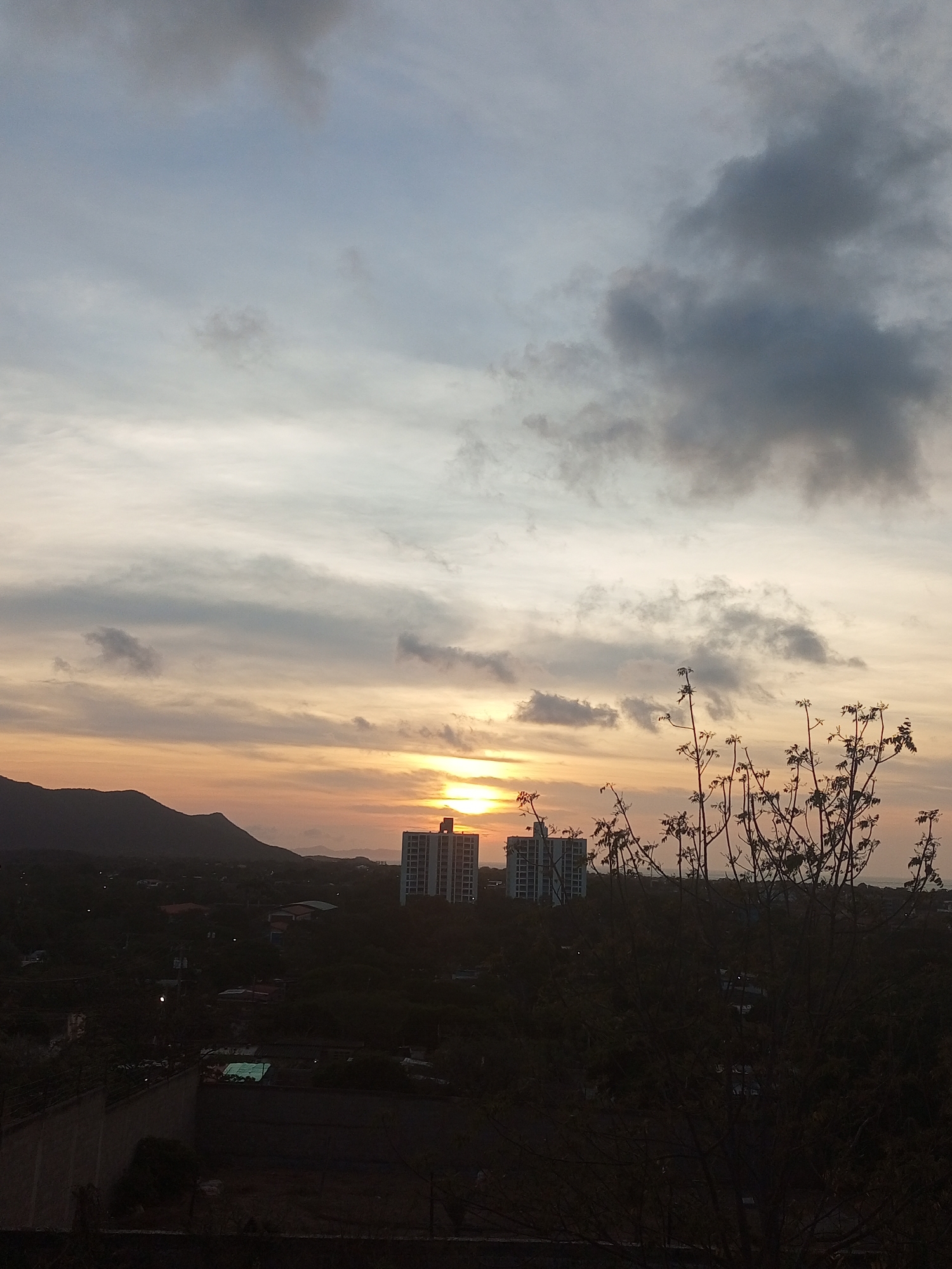 A wide-angle shot captures a sunset over the urban area of Juan Griego, Venezuela. The skyline is punctuated by two high-rise buildings silhouetted against the setting sun. A mountain range is faintly visible in the distance to the left. In the foreground, a partially obscured leafless tree adds a sense of depth to the scene. The foreground is dark, with a wall and vegetation adding a sense of enclosure. The sky is filled with layered clouds tinged with orange hues from the sunset, while darker clouds loom overhead.