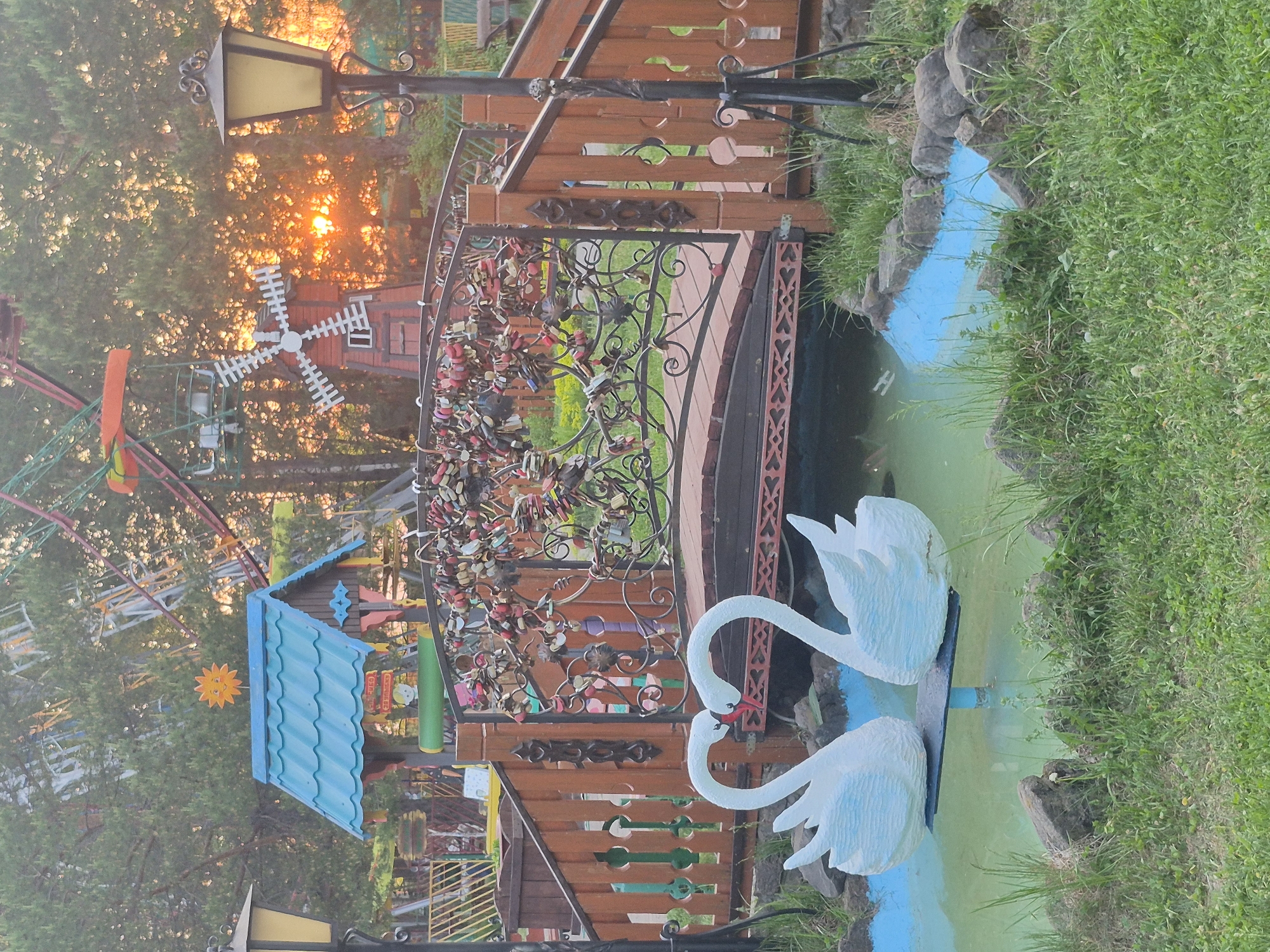 The image captures a whimsical park scene in the city of Unknown, Unknown. In the foreground, two white swan statues with red beaks face each other in a pond painted with blue and green hues. The edges of the pond are lined with green grass. A small wooden bridge with a black iron railing heavily adorned with padlocks spans the pond. The bridge is decorated with cut-out shapes.
In the background, the sun sets amidst trees, casting a warm glow. Structures from an amusement park, including a Ferris wheel, a windmill, and small buildings with brightly colored roofs, are visible. A black lamppost is positioned near the bridge, with its light fixture facing away from the viewer. The scene is colorful and playful, suggesting a recreational or tourist attraction.