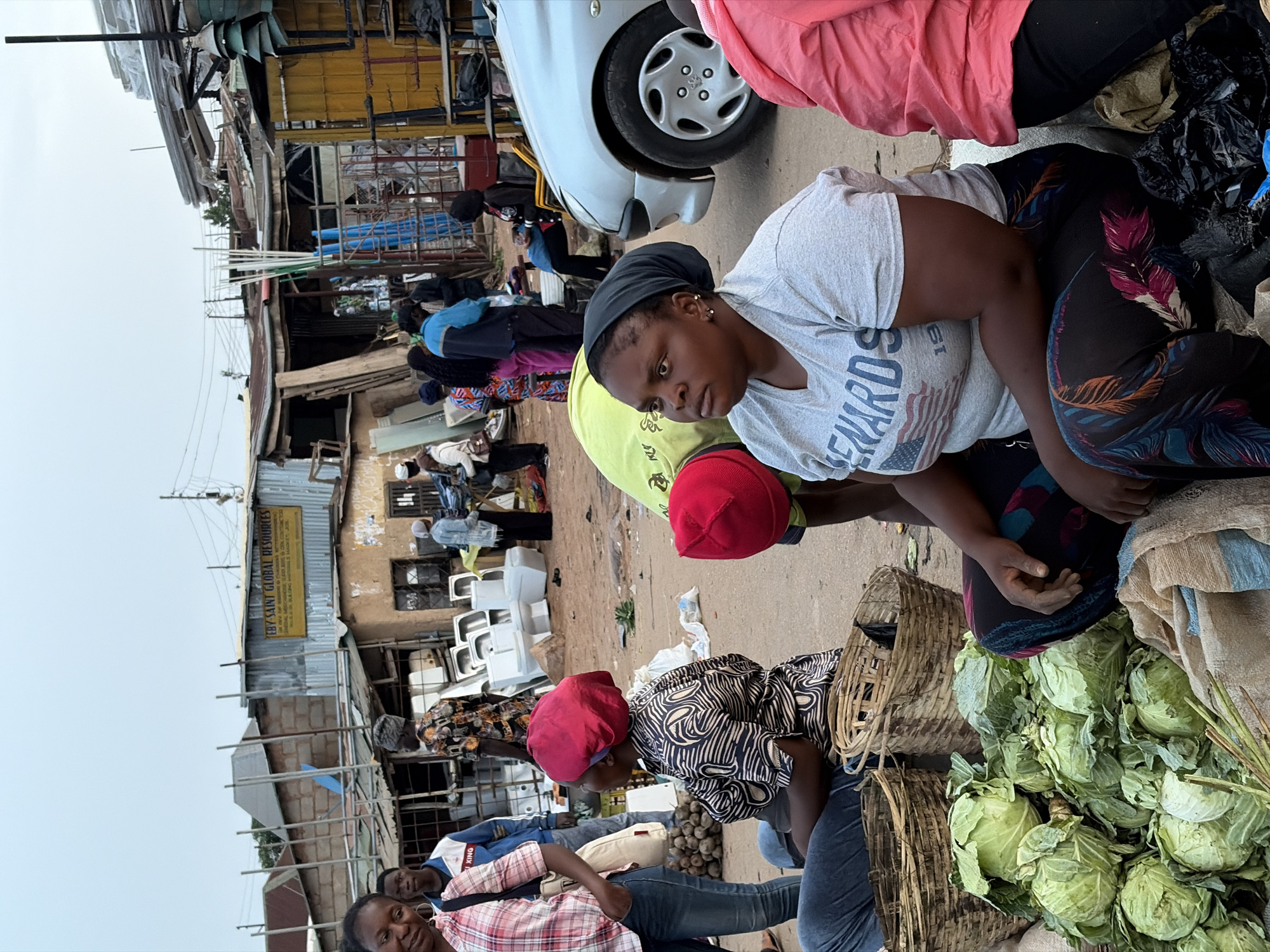 The image depicts an outdoor market scene in Gura Top, Nigeria. In the foreground, two women are seated near baskets and a pile of cabbages, appearing to be engaged in commerce. The woman in the center wears a light-gray t-shirt, a black headscarf, and patterned pants, while the woman next to her wears a patterned top and a red cap. To the left, another woman in a plaid shirt and jeans is seated. Behind them, there are more people further back, some also appearing to be seated and potentially part of the market. A silver car is partially visible in the upper-left portion of the image. In the background are buildings, some appearing to be commercial establishments, with signs written in English. The sky above is overcast.