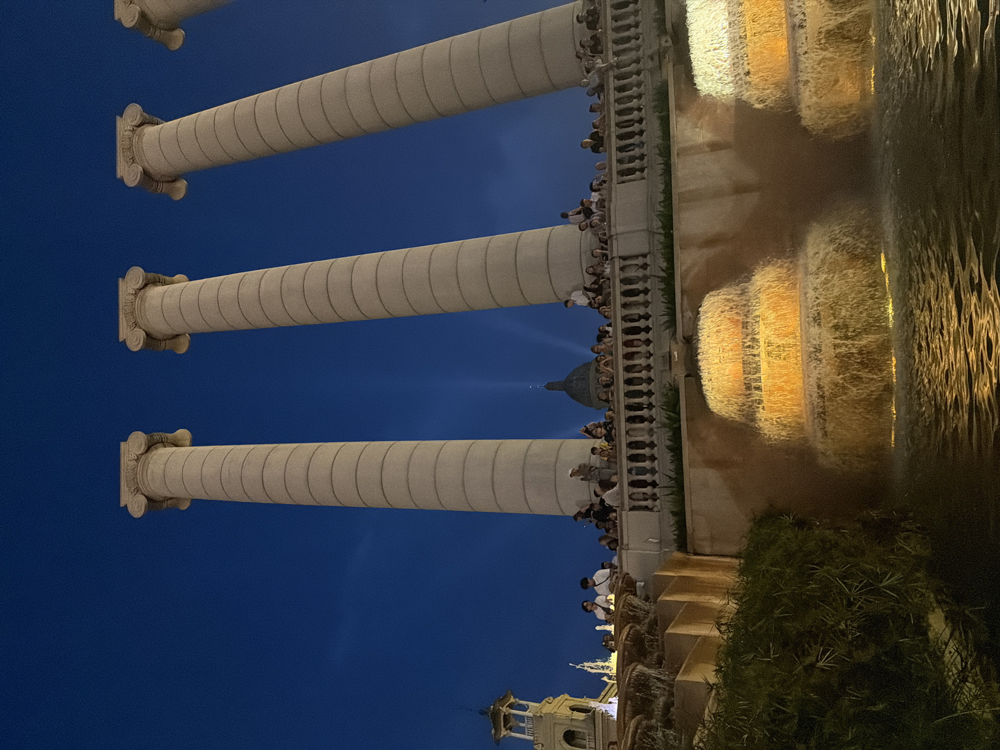 This image captures a nighttime view of the Magic Fountain of Montjuïc in Barcelona, Spain. Four large, fluted columns, characteristic of classical architecture, dominate the upper portion of the frame, set against a deep blue night sky. Below the columns, a balustrade with decorative posts lines a raised walkway where a crowd of people is gathered, observing the scene. In the mid-ground, a tiered fountain is illuminated with warm, golden light, creating a cascading waterfall effect. The water from the fountain flows into a pool in the foreground, reflecting the lights and the surrounding structures. To the left, a tower with an ornate roof and arches is visible. In the background, a dome and a faint light beam suggest other architectural features of the area. The overall atmosphere is one of public gathering and enjoyment of a nighttime spectacle.