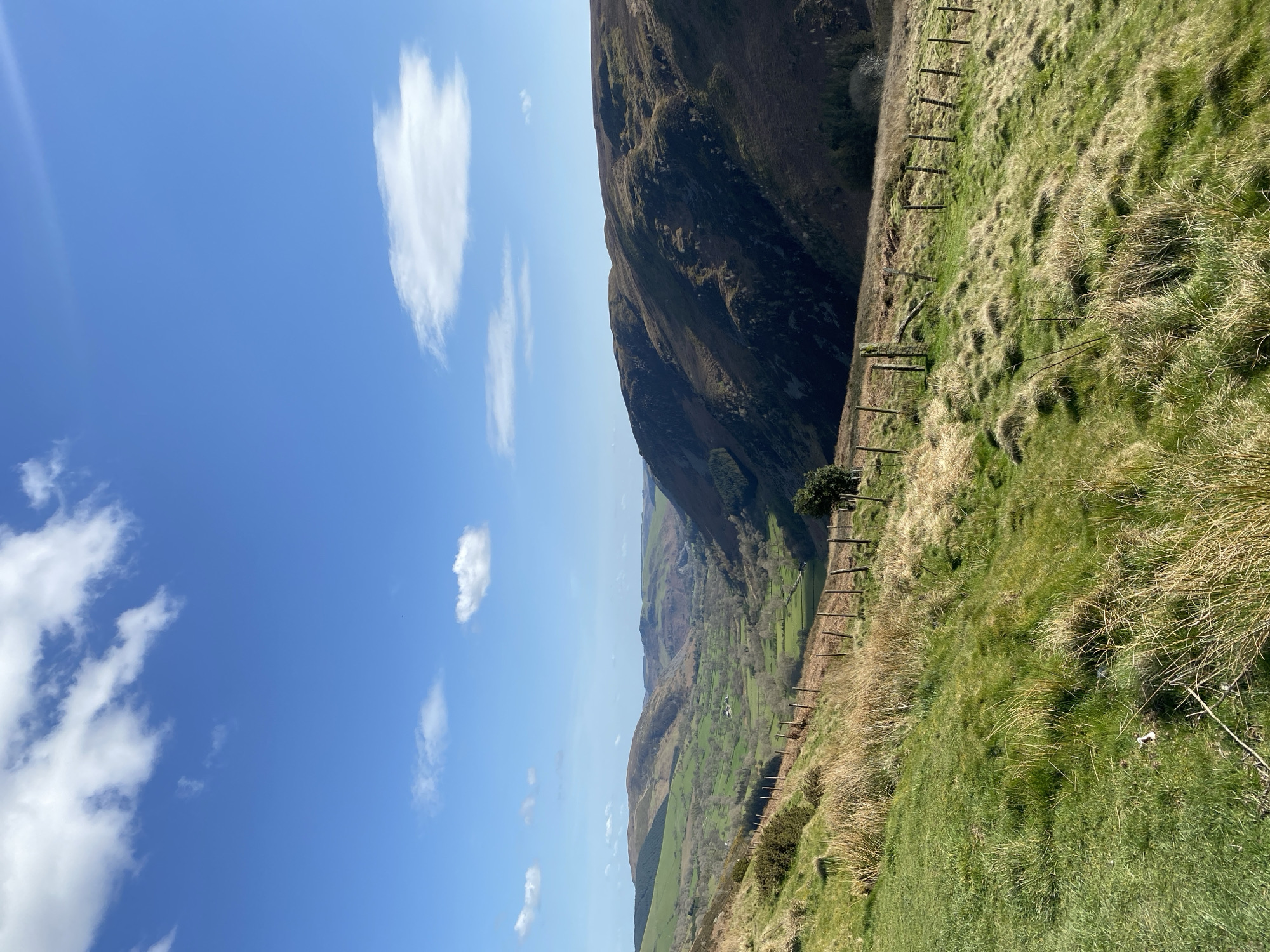 The image shows a rural scene near Llangynog, United Kingdom. A grassy hillside occupies the foreground and right side of the image. A wooden fence runs along the slope. Beyond the fence, a valley is visible with green fields and scattered trees. A dark, steep mountain rises to the left. The sky is blue with scattered white clouds. The image is oriented vertically.