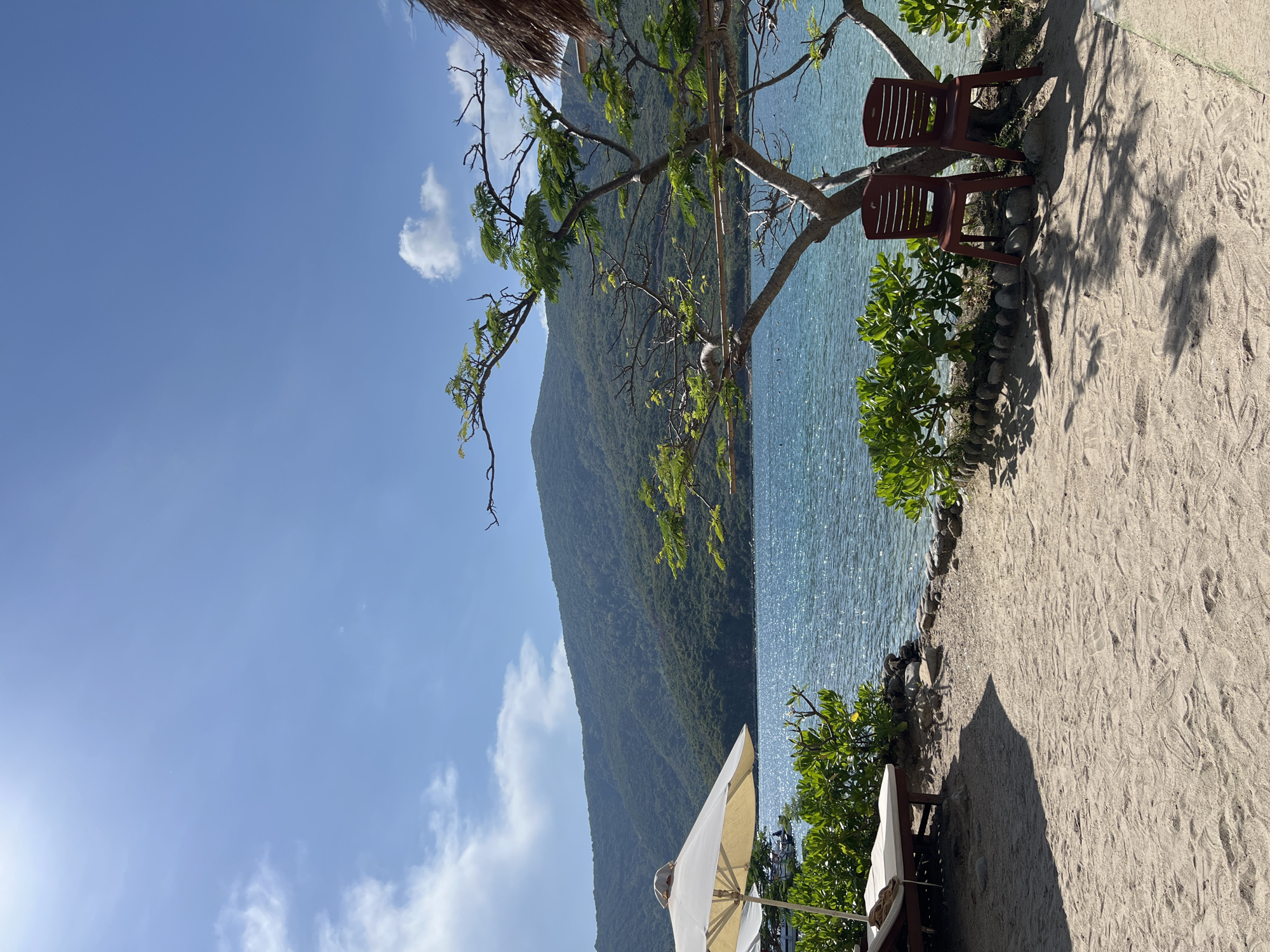 The image captures a scenic view of a beachside location in Nha Trang Ward, Vietnam. The scene showcases a beach with light-colored sand, bordering blue water. A large, green mountain rises in the background. In the foreground, there are two simple, maroon plastic chairs positioned along the shoreline. A small tree with sparse green leaves overhangs the seating area. A white umbrella stands partially visible in the lower section of the frame. The sky above is mostly blue, with a few scattered white clouds. The light and shadows suggest a sunny day.