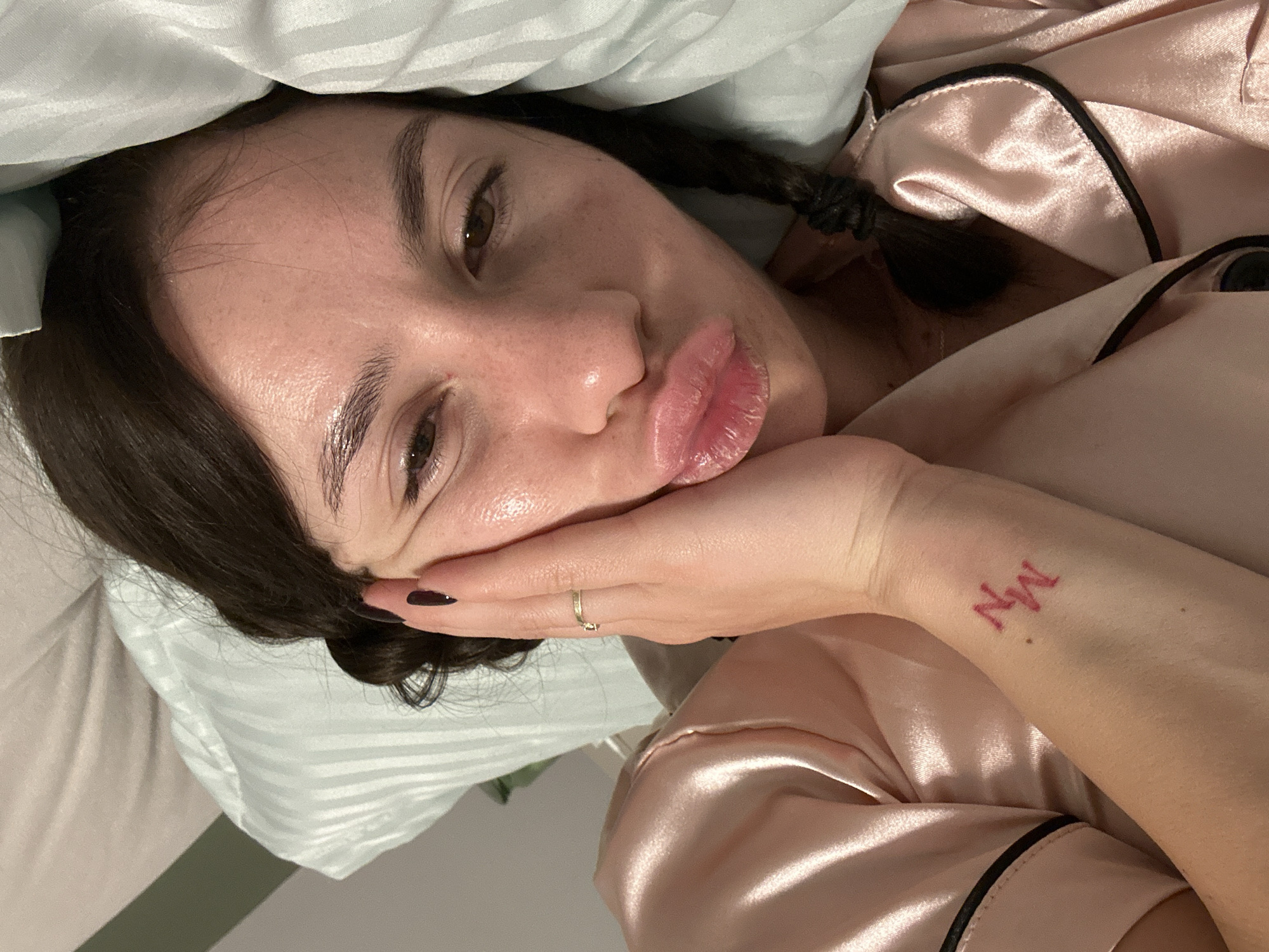 A woman is lying in bed, possibly indoors. She has dark hair in braids and is wearing pink pajamas. She has a tattoo on her wrist with the letters "NM". The environment consists of a bed with a pillow. The location is not discernible from the image.