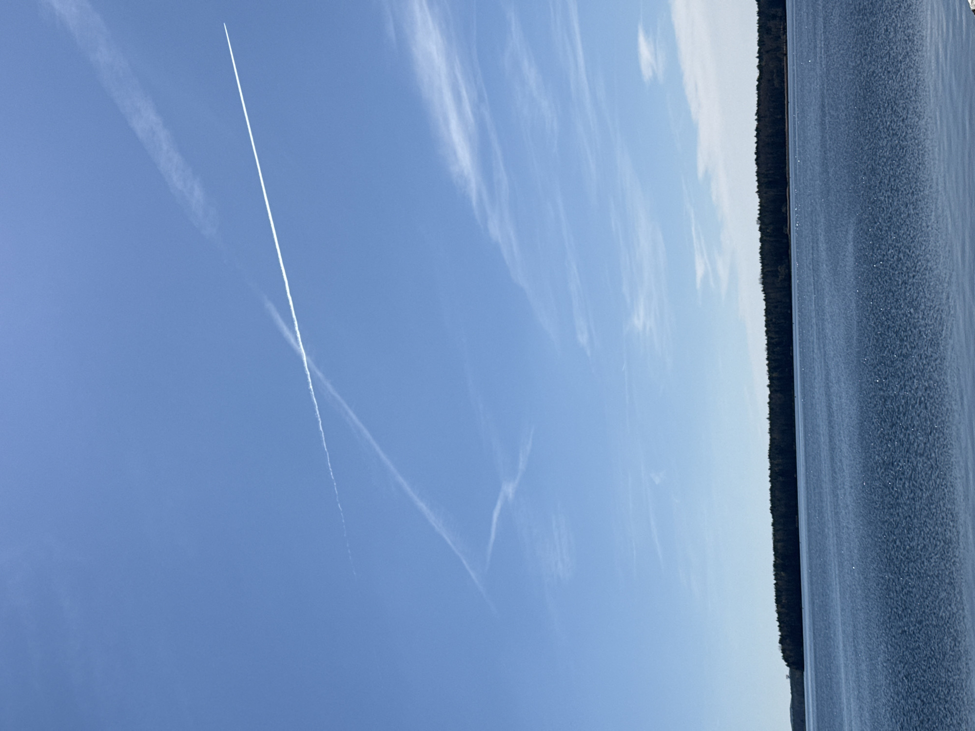 The photograph depicts a landscape scene in Senftenberg - Zły Komorow, Germany. The sky is a clear blue with contrails from aircraft. Wispy clouds are scattered throughout. On the right side of the frame is a body of water with a dark shoreline densely populated with trees.