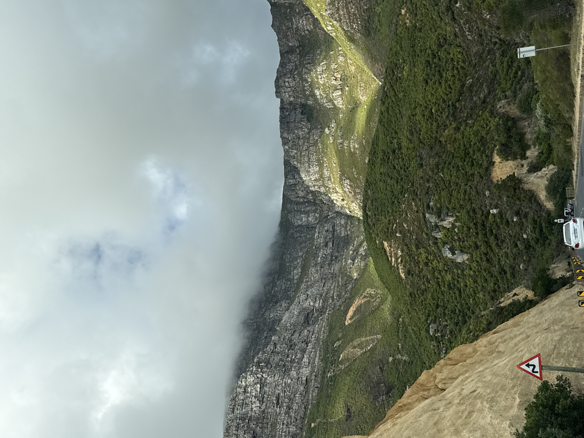 The image presents a landscape scene in Hout Bay, South Africa. A large, steep mountain with vertical rock formations dominates the view, its left side obscured by low-lying clouds. Green vegetation covers the right side of the mountain. The lower right corner of the image includes a road with a vehicle, with a road sign indicating a series of curves or bends ahead. Below the sign, there is a steep drop.