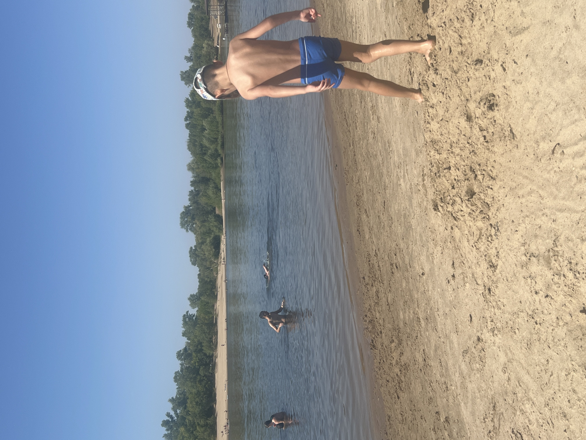 A medium shot shows a sandy beach with a body of water and a tree-lined shore in the background. The sky is clear and blue. In the foreground, a young boy wearing blue swim trunks and a patterned cap faces away from the camera, his back to the viewer. He is standing on the sand, and his skin appears to be wet and shiny, suggesting he may have recently been in the water. Further out in the water, several other individuals are visible. One person is swimming, and two others are standing in the water. The beach is lightly populated, with a few scattered figures and loungers visible along the shore in the distance. The environment suggests a sunny, recreational day at a beach, possibly on the Volga River near Kilyakovka, Russia.