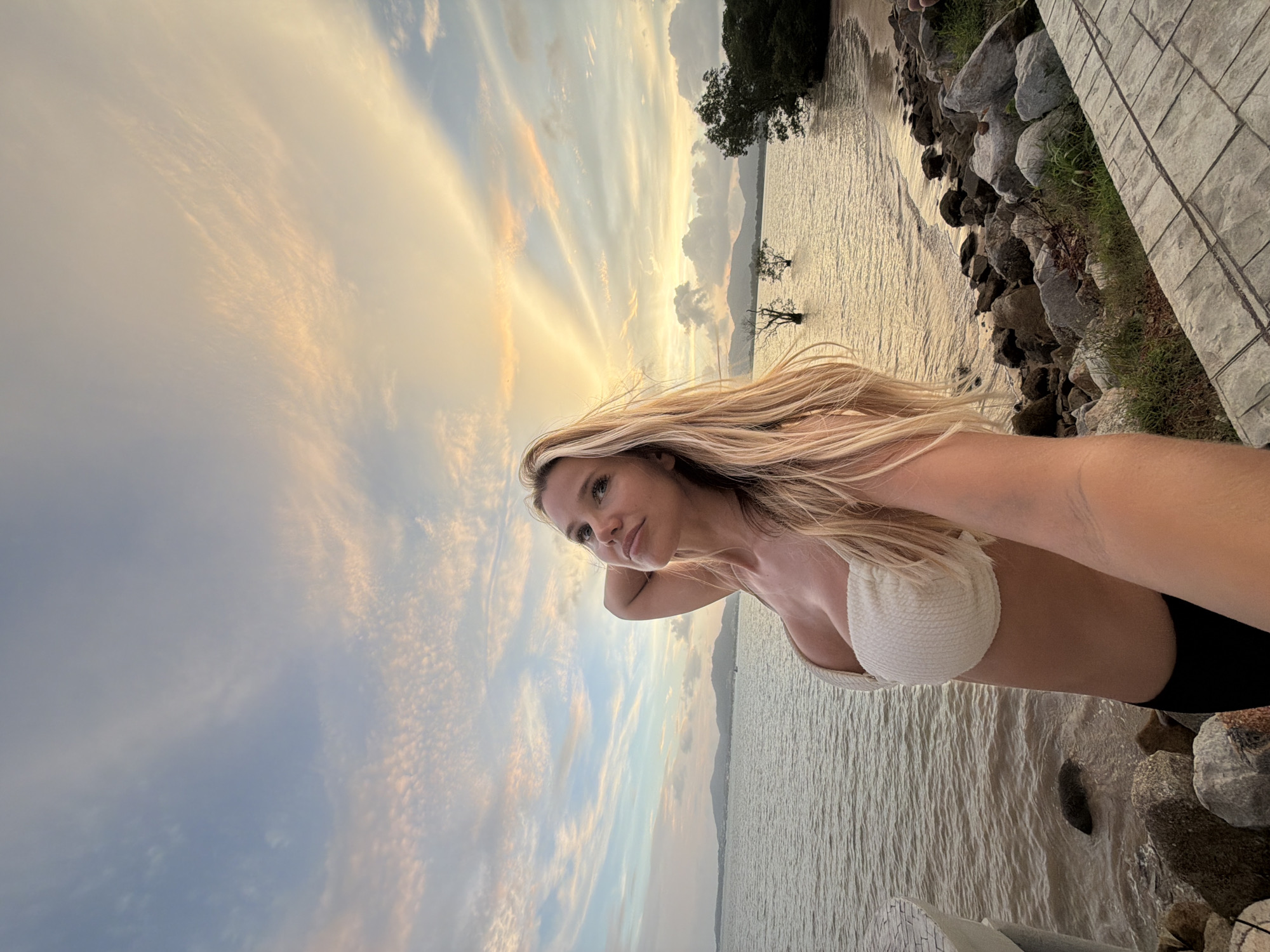 A blonde woman in a white bikini top stands on a rocky shore, taking a selfie. She is looking up and to the left, with her right arm raised behind her head. The ocean is to her left, and a beach with trees is to her right. The sky is filled with clouds, and the sun is setting. The location is the city of Chalong, Thailand.