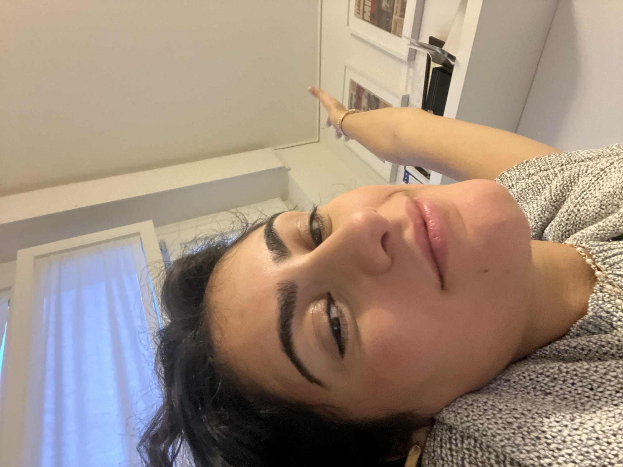 A young woman, appearing to be of Middle Eastern or Mediterranean descent, is captured in a close-up selfie, lying down with her head tilted and looking directly at the camera. She has dark, wavy hair, prominent eyebrows, and full lips, and is wearing a light grey, textured sweater. On her extended left wrist, she wears a gold-toned bracelet with white beads, and a small gold earring is visible on her left earlobe. Her left arm is raised, with her index finger pointing upwards.
The setting is an indoor room in Milan, Italy. To the left side of the frame, a window with white, semi-sheer curtains allows soft light to filter in, creating a slightly cool tone. To the right, a white wall features shelves displaying several framed items, which appear to be newspaper clippings or prints, consistent with European publications. A dark, rectangular object, possibly a book or electronic device, is also on a shelf. The ceiling above is plain white. The woman's expression is neutral and calm.