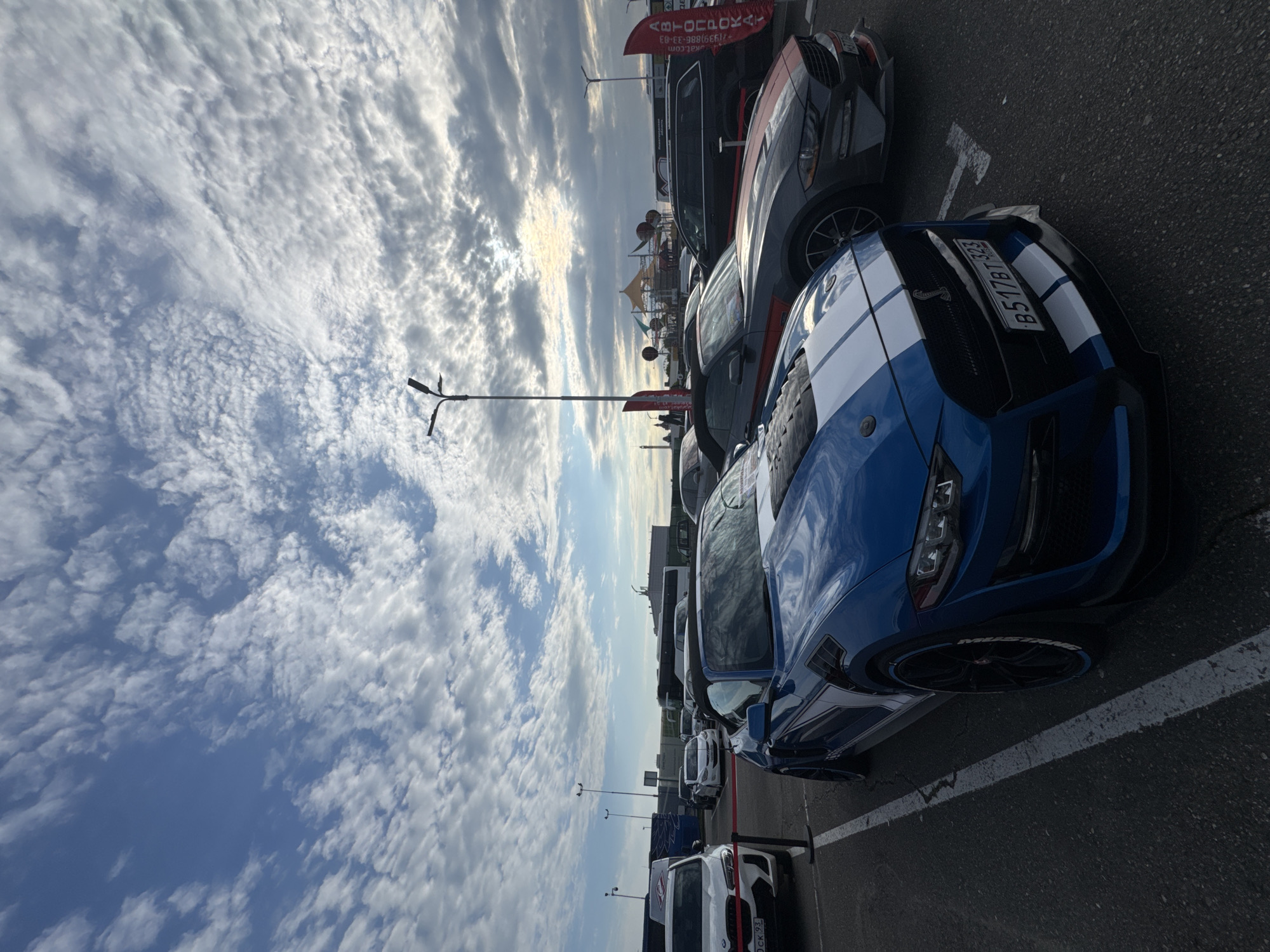 The image depicts a parking lot in Sirius, Russia, under a partly cloudy sky. The focus is on a row of parked cars, with a blue Ford Mustang with white racing stripes prominently featured in the foreground. The Mustang has a license plate with the number "B517BT 323". Behind the Mustang are several other vehicles, including a gray car and a white car. A few light poles are visible in the background along with some banners. Beyond the cars, there is a structure with colorful awnings. The parking lot is marked with white lines, and the asphalt surface is visible.