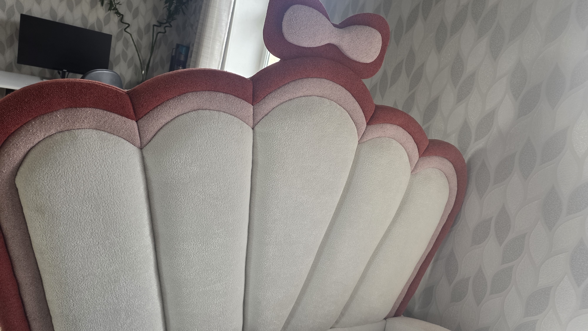 The image shows a close-up view of the back of a decorative chair with a shell-like design and a bow on top. The chair is upholstered in textured fabric in shades of red, pink, and light grey. The background features a patterned wallpaper in shades of grey and white, with vertical blinds visible on the right side of the frame. A desk with a black monitor is visible in the upper left corner, along with a plant in a vase and a shelf with some items. The scene appears to be in an indoor setting, likely a room in a home or office. The location in the city of Chuy, Kyrgyzstan, is not discernible from the visual information provided in the image.