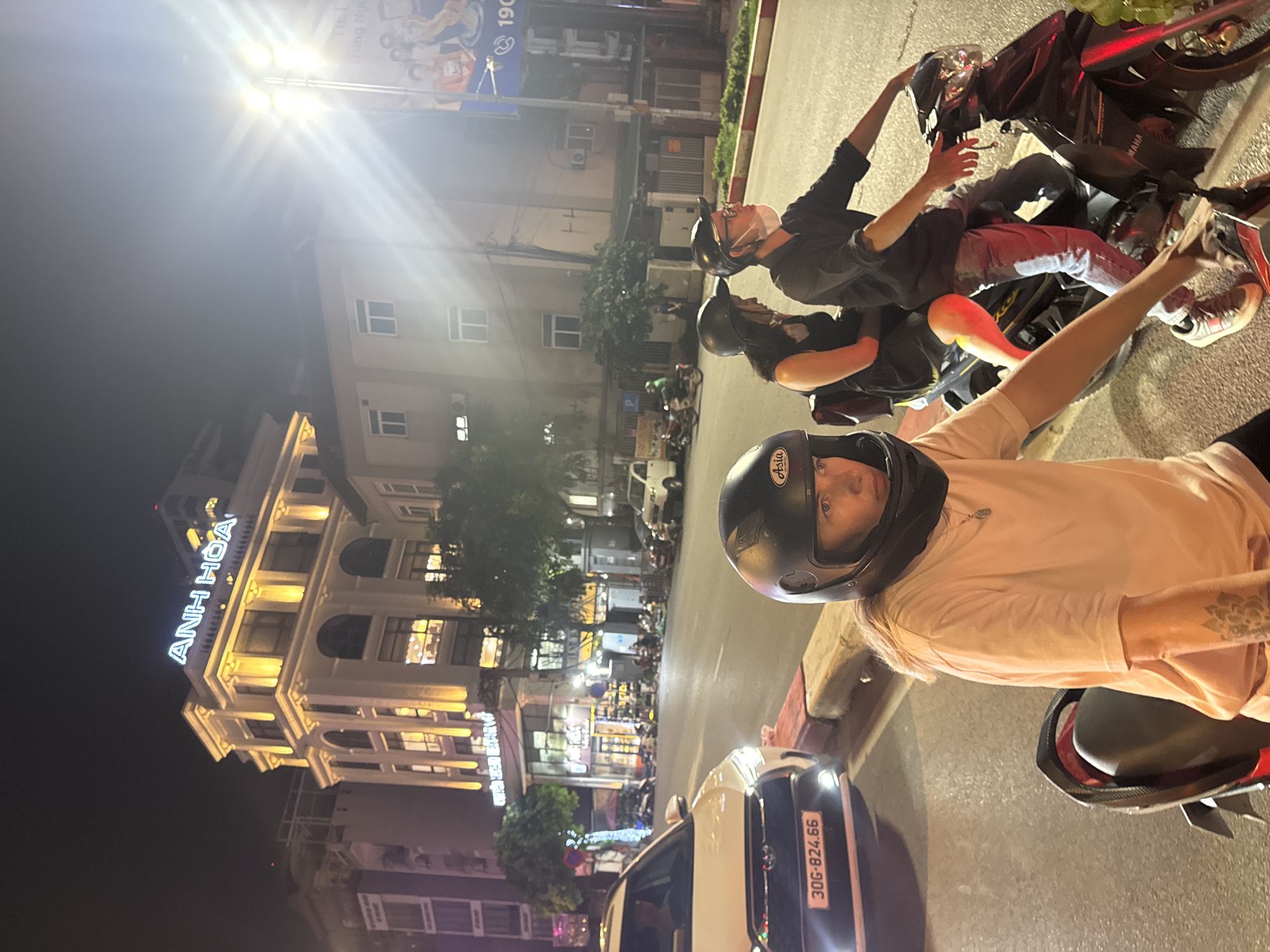 In Hà Nội, Vietnam, at night, a white Toyota Cross with license plate "30G-824.66" is parked on the right side of the road. In the foreground, a person wearing a light pink t-shirt and a black helmet is sitting on a motorcycle. To their right, a couple on another motorcycle is also visible. The man is wearing a black shirt and a face mask, and the woman is wearing a black dress. Behind them, a brightly lit building displays the sign "ANH HOA". The street is illuminated by streetlights and the lights from the building. Other vehicles and motorcycles are present on the street, suggesting a busy urban environment.