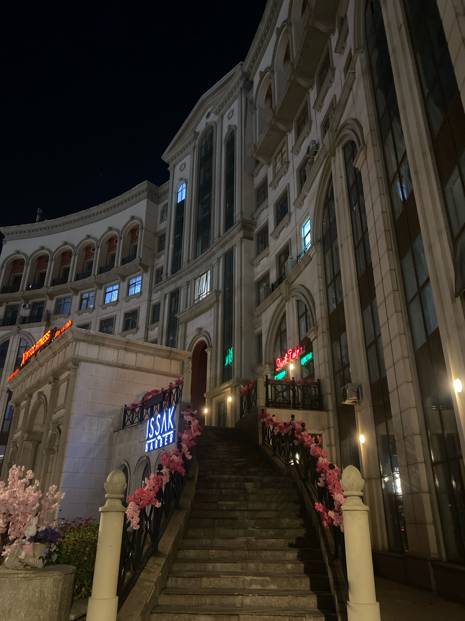 The image shows the exterior of a multi-story building at night, located in Astana, Kazakhstan. The building has a classical architectural style with arched windows and decorative elements. A wide stone staircase with decorative railings adorned with pink artificial flowers leads up to an entrance. Above the entrance, there are neon signs. One sign reads "ISSAK BARBER" in blue neon letters. Another neon sign on the left side of the building appears to read "XFITNESS CLUB" in red neon letters. The surrounding environment is dark, indicating it is nighttime. There are no people visible in the scene. The lighting comes from the building's illuminated windows and the neon signs.