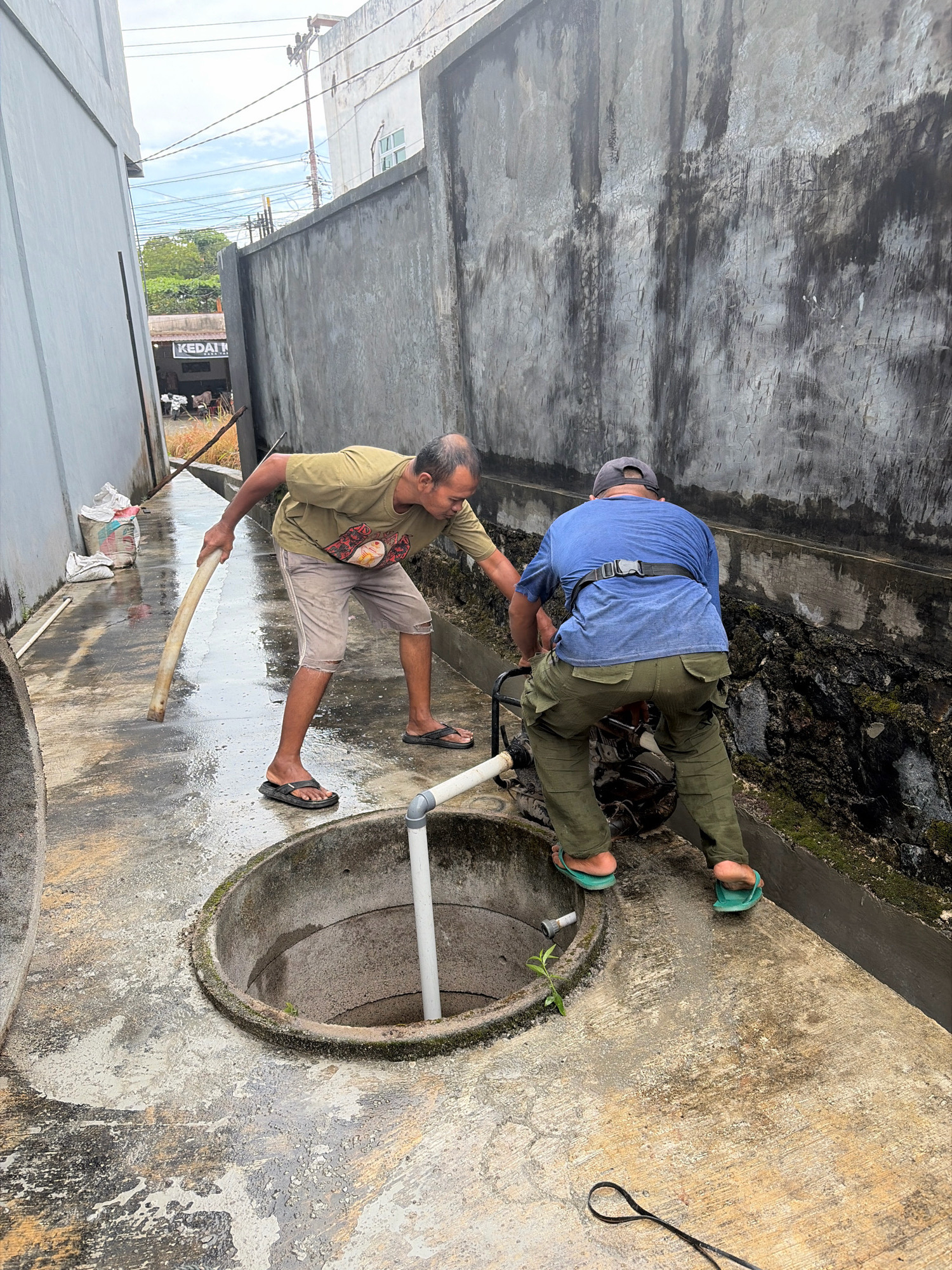 Two men are working near a circular concrete opening in the ground, likely a well or manhole, in what appears to be a narrow alleyway in Tanjung Pinang, Indonesia. The alley is paved with concrete, wet and reflective, suggesting recent rain or water usage. On the left, a man in a light olive t-shirt and khaki shorts is using a long, curved wooden pole to push or pull something into the opening. He is wearing dark flip-flops. On the right, a man in a blue t-shirt, green cargo pants, and a grey baseball cap is operating a piece of machinery, possibly a pump, positioned near the opening. He is wearing green flip-flops. The walls of the alley are made of rough, weathered concrete. One wall is light gray, and the other is dark gray with vertical streaks and cracked texture. In the background, there are power lines and some greenery visible, along with a sign that reads "KEDAI." Bags of what appear to be cement or sand are stacked against the light gray wall on the left. The scene suggests manual labor or maintenance work being performed in an urban environment.