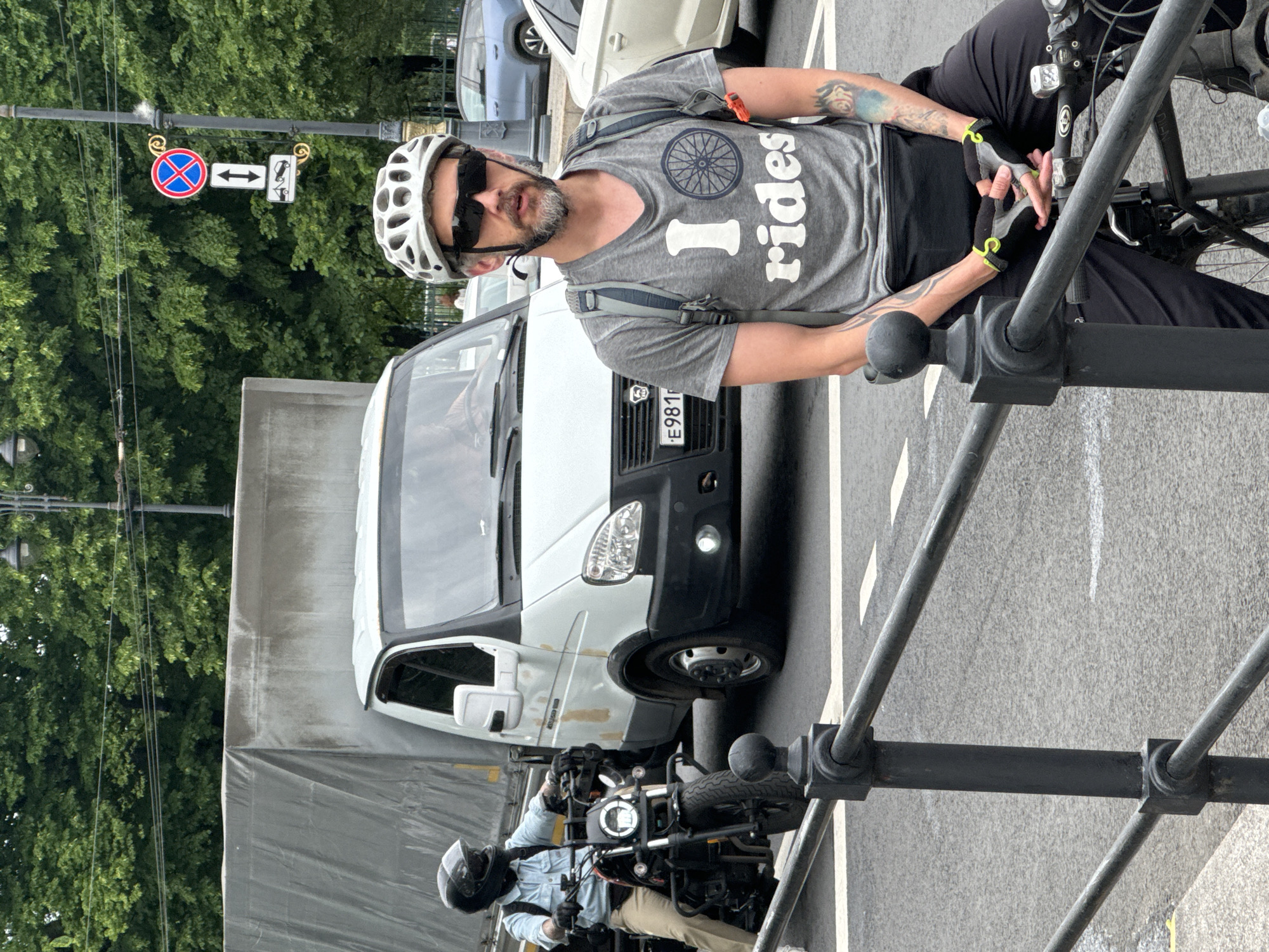 The image depicts a street scene in Saint Petersburg, Russia. In the foreground, a man wearing a grey t-shirt with the slogan "I rides" and a bicycle helmet is seated on a bicycle. He has a beard and sunglasses and is wearing black pants and cycling gloves. His arms are adorned with tattoos. Behind him, a white van is parked on the street, and to the left, another person on a motorcycle is visible. In the background, trees and a lamppost with traffic signs are present. The scene appears to be a public street with asphalt paving and a metal railing in the foreground.